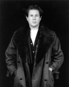 The artist Julian Schnabel from the hips up facing and looking straight at the viewer. His hands are in the pockets of his trench coat with dark shearling lapels.