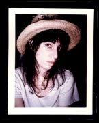 Close up of a woman in a white t-shirt and straw hat with black hair looking into the camera.