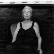 The artist Alice Neel sitting facing the viewer in a black dress in front of a black brick wall with white molding. Her eyes are looking slightly to the side of the camera.