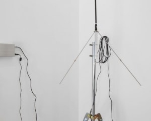 A tight photograph of the sound technology associated with the artwork. The silver transmitter installed upon the wall is at right, and the antennae is tight in the foreground.