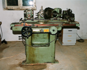 A photograph of a grinder from a factory, seafoam green with oil stains on it, sitting in a desolate room with gray walls and one window