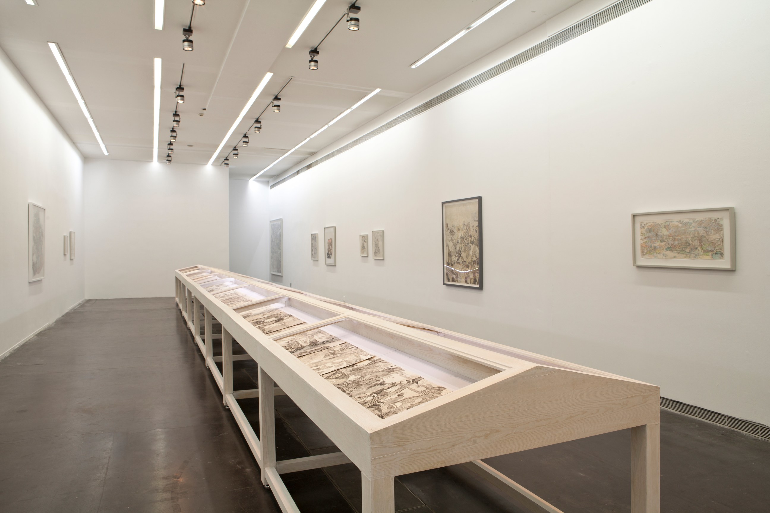 A gallery space with ink drawings installed on walls and a wooden display table placed at the center