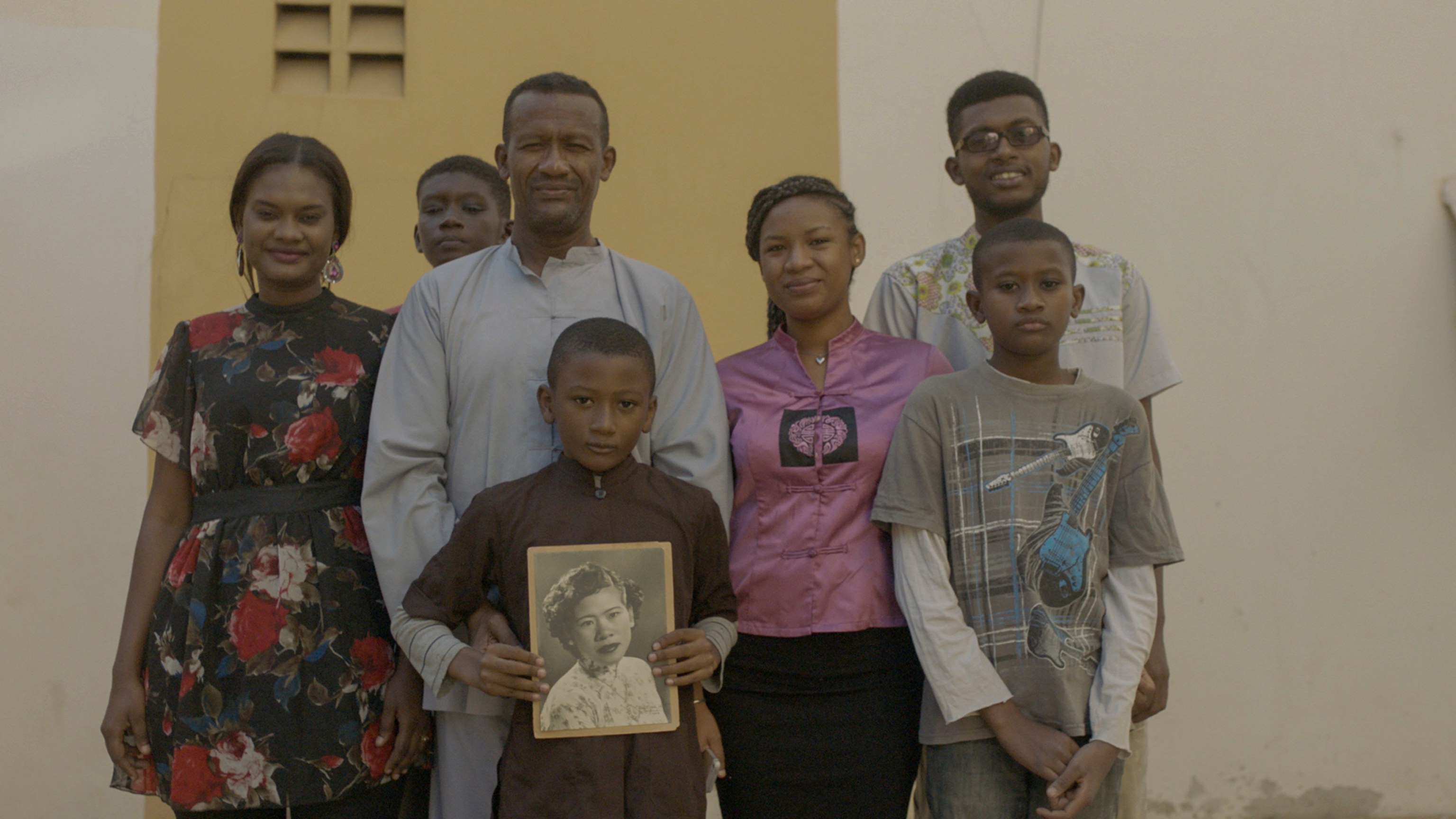 Video still of a family, with a young boy holding up a black and white photo in The Specter of Ancestors Becoming, 2019
