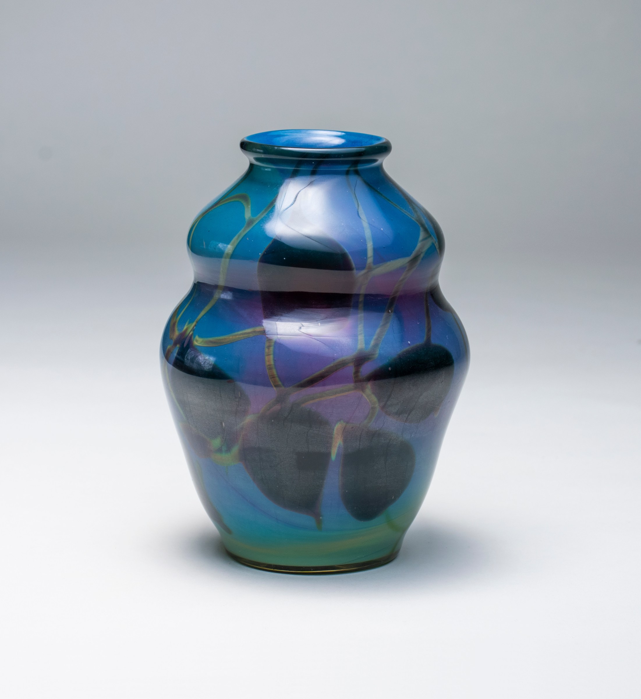 A tiffany favrile glass reactive paperweight style vase, the blue double gourd shaped body shifting in color and opacity as you move around the vase, decorated with all over motif of swirling vines and heart shaped leaves in a dark green glass. The vase appears to glow purple in transmitted light.