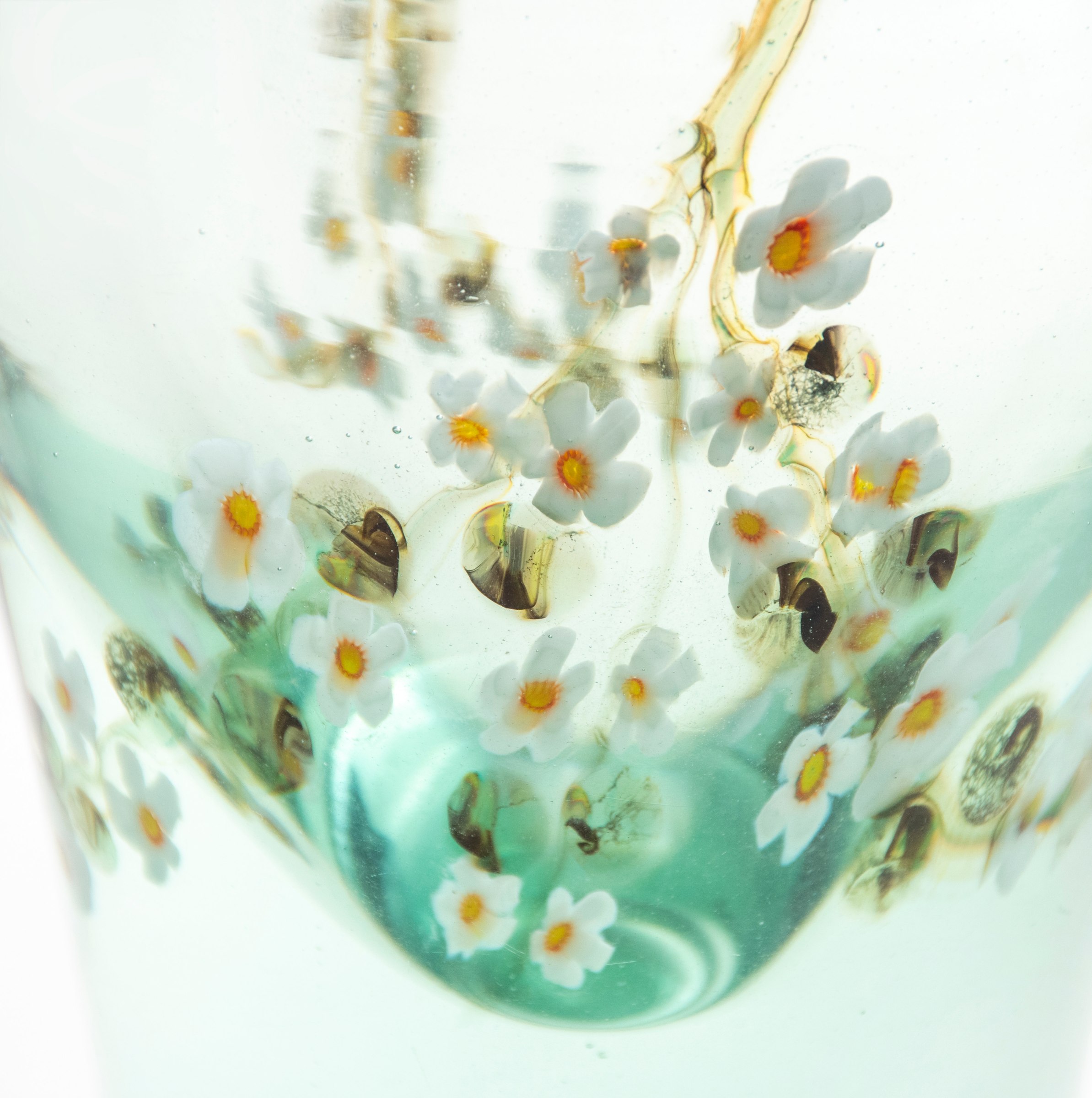 A close up view of the encased design of an apple blossom branch with many white flowers, inside a thick layer of clear glass tinted green to mimic a pool of water, in a tiffany favrile glass aquamarine vase.