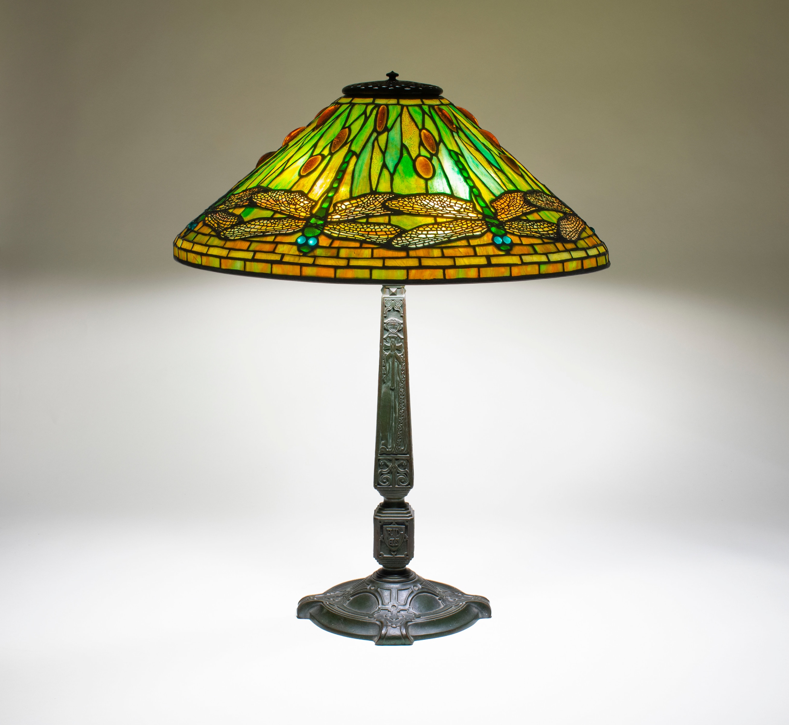 an iconic tiffany dragonfly lamp, the shade with green background glass shifting in tones of orange and yellow, the dragonflies with kelly green bodies and teal eyes with orange wings overlaid with openwork metal filigree to represent the insect