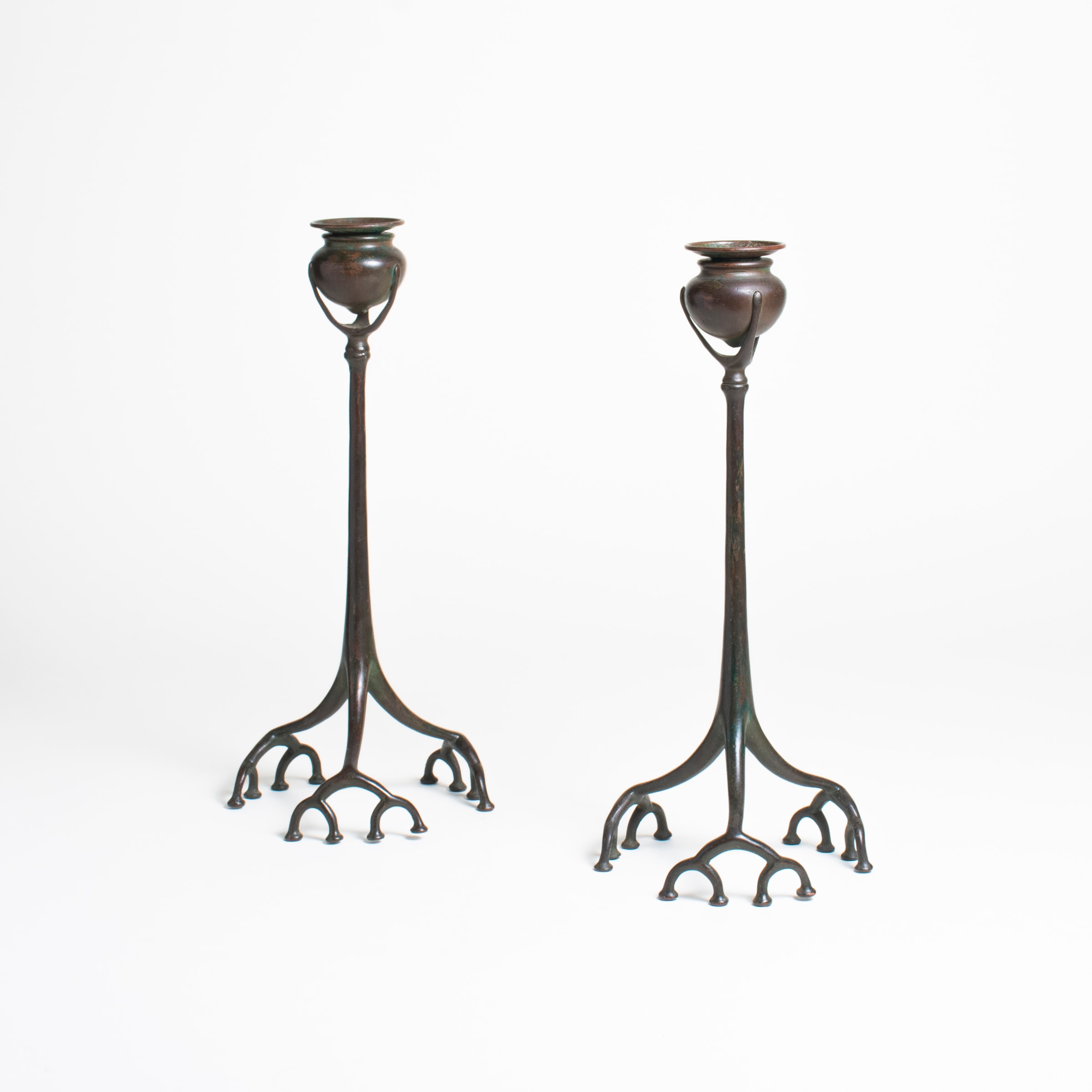 a pair of bronze candlesticks by tiffany studios, the thin bronze stems supporting rounded candle cups, the bases formed by three &quot;feet&quot; of fractal-like root structures in thin bronze