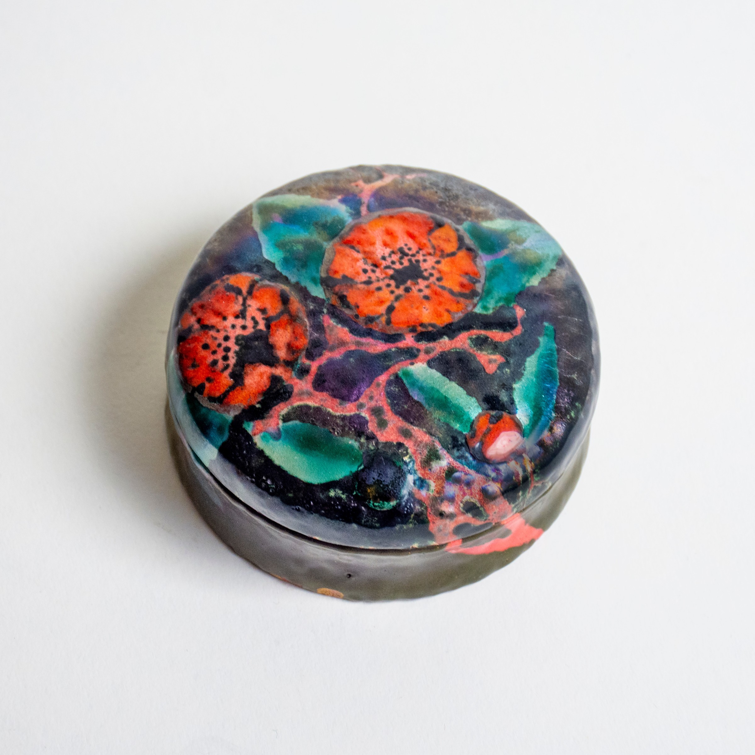 a rare example of tiffany metalwork, a small circular lidded trinket box, the background a dark black/green color, with vivid decoration of bright red poppies and green leaves on the surface of the lid.