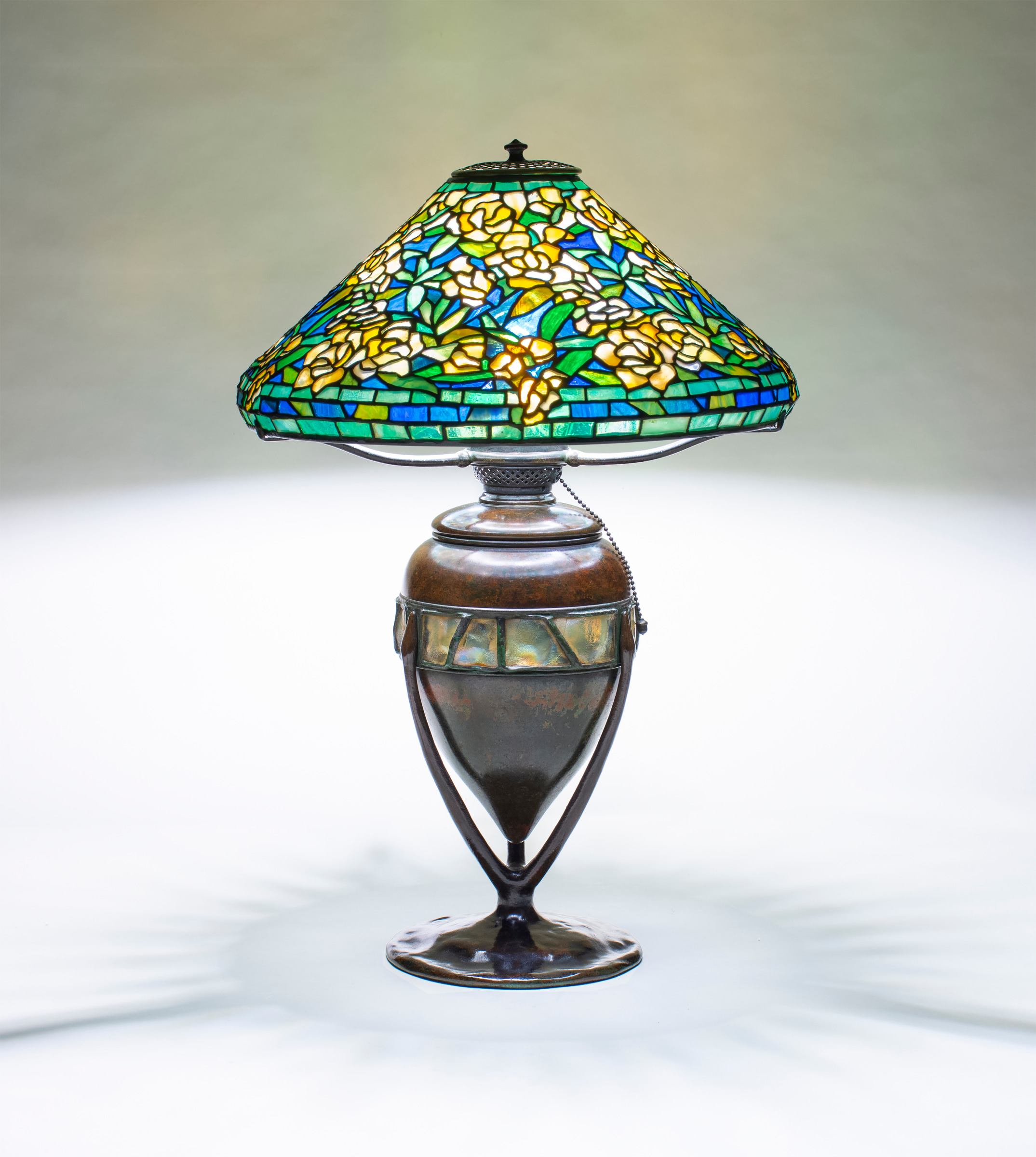 A Tiffany Studios Lamp, with a Wild Rose pattern shade with mottled yellow flowers against a deep blue sky, paired with a bronze table base in brown patina, the body with a horizontal row of inset Tiffany Favrile Glass Turtle Back Tiles with uneven gold iridescent surfaces.