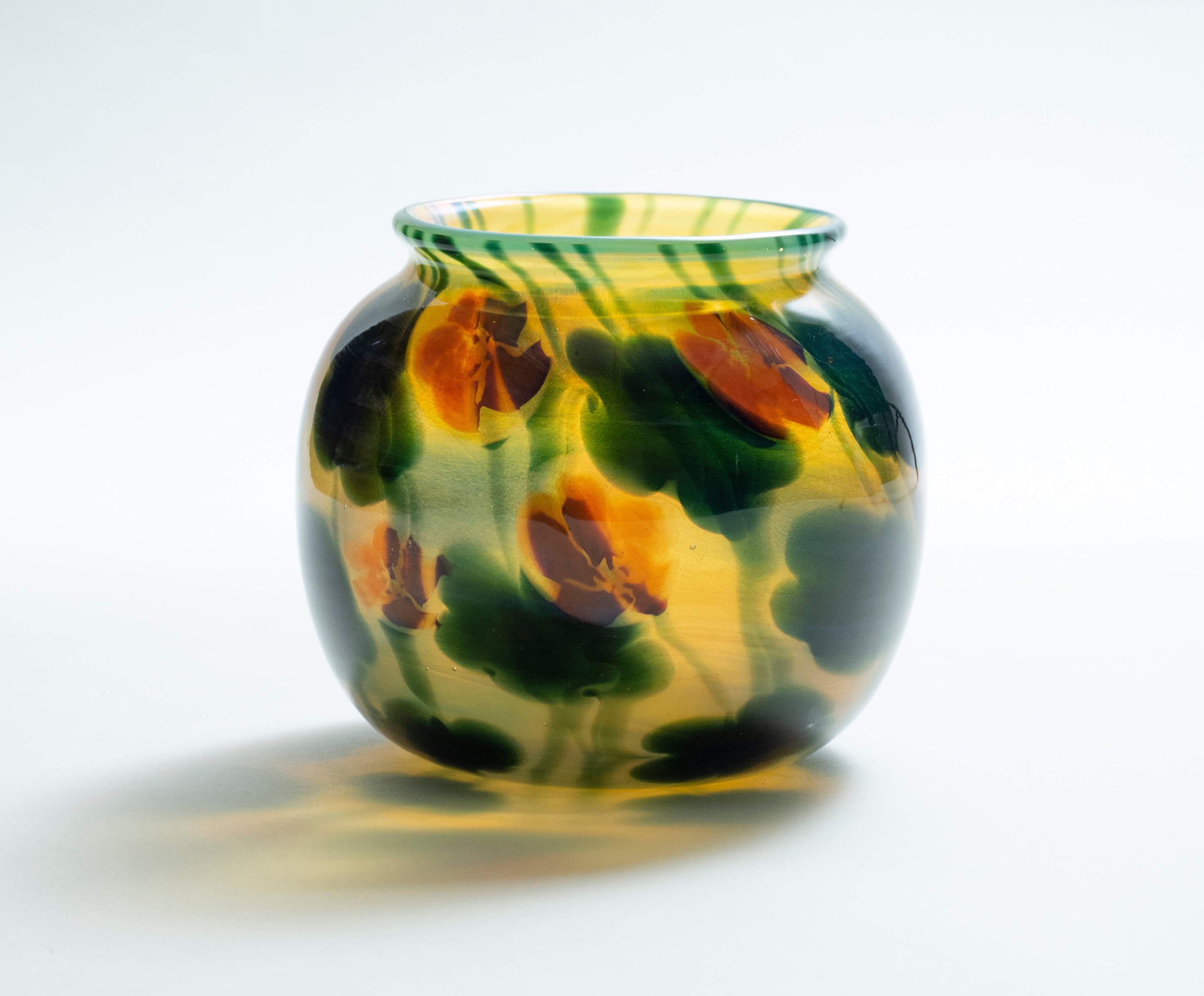 A blown Tiffany favrile glass paperweight style vase, a wide round body, the background transparent golden yellow with swirling thin vines and round nasturtium leaves, interspersed with bright red nasturtium flowers.