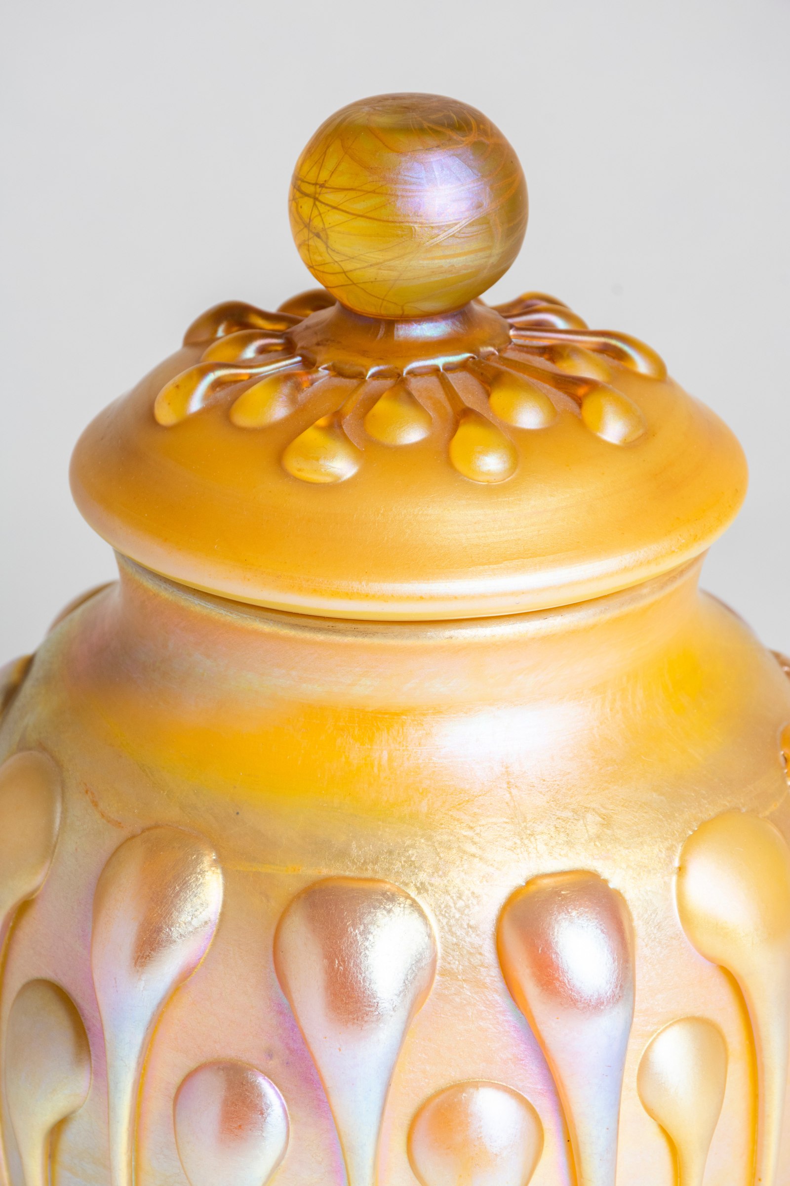 A close up photograph showing the texture of the gold iridescent applied drops on both the vase body and the matching lid in addition to the round glass finial which is swirled with shades of yellow.