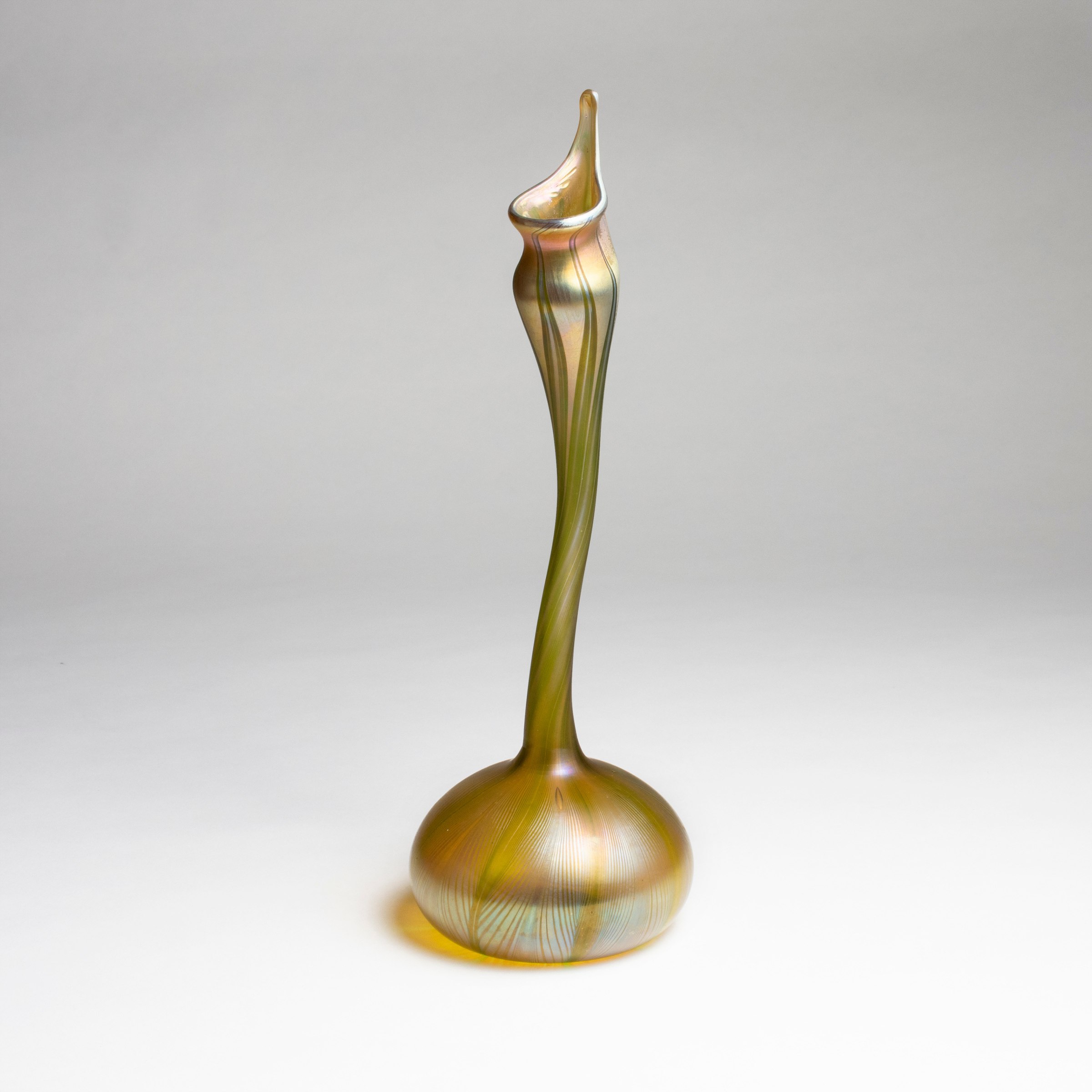 an early piece of blown tiffany glass with a rounded foot, rising to an irregular wavy thin stem, known as a gooseneck vase or rosewater sprinkler, gold iridescent glass decorated with a pulled feather motif