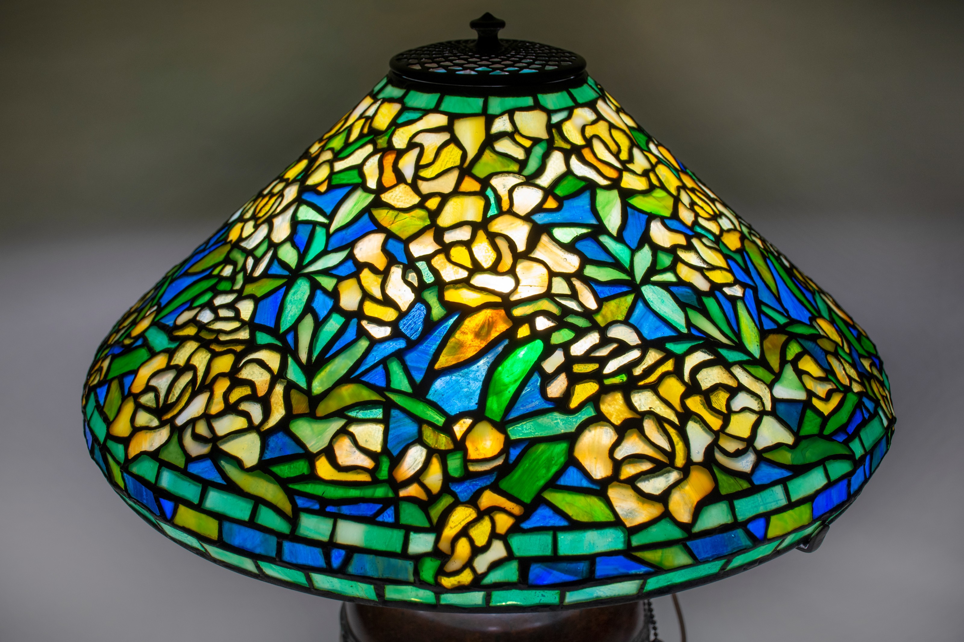 a close up showing a leaded glass Tiffany lamp shade in the complex and early &quot;Wild Rose&quot; pattern; this example has many small roses formed by tiny pieces of glass in a bright yellow with mottling. The leaves are vivid green, the background glass an even deep blue.