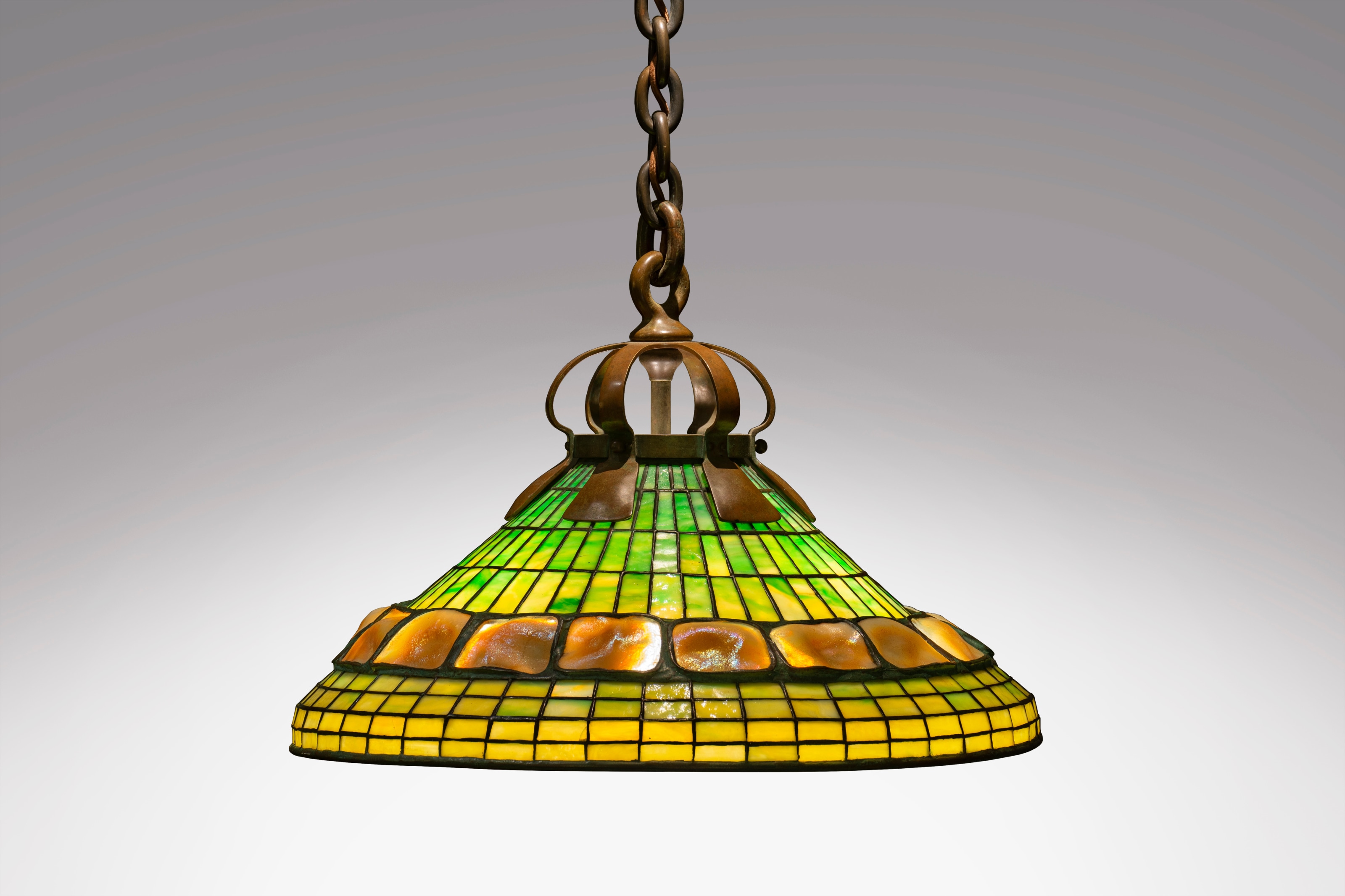 A hanging Tiffany Studios Lamp Shade, hanging from a thick bronze chain with heavy links, the shade suspended from thick arts &amp; crafts style flattened bronze ribs, the leaded glass cone shaped-shade in light green near the crown and shifting to yellow near the lower rim, with an inset horizontal band of gold iridescent molded tiffany glass turtle back tiles