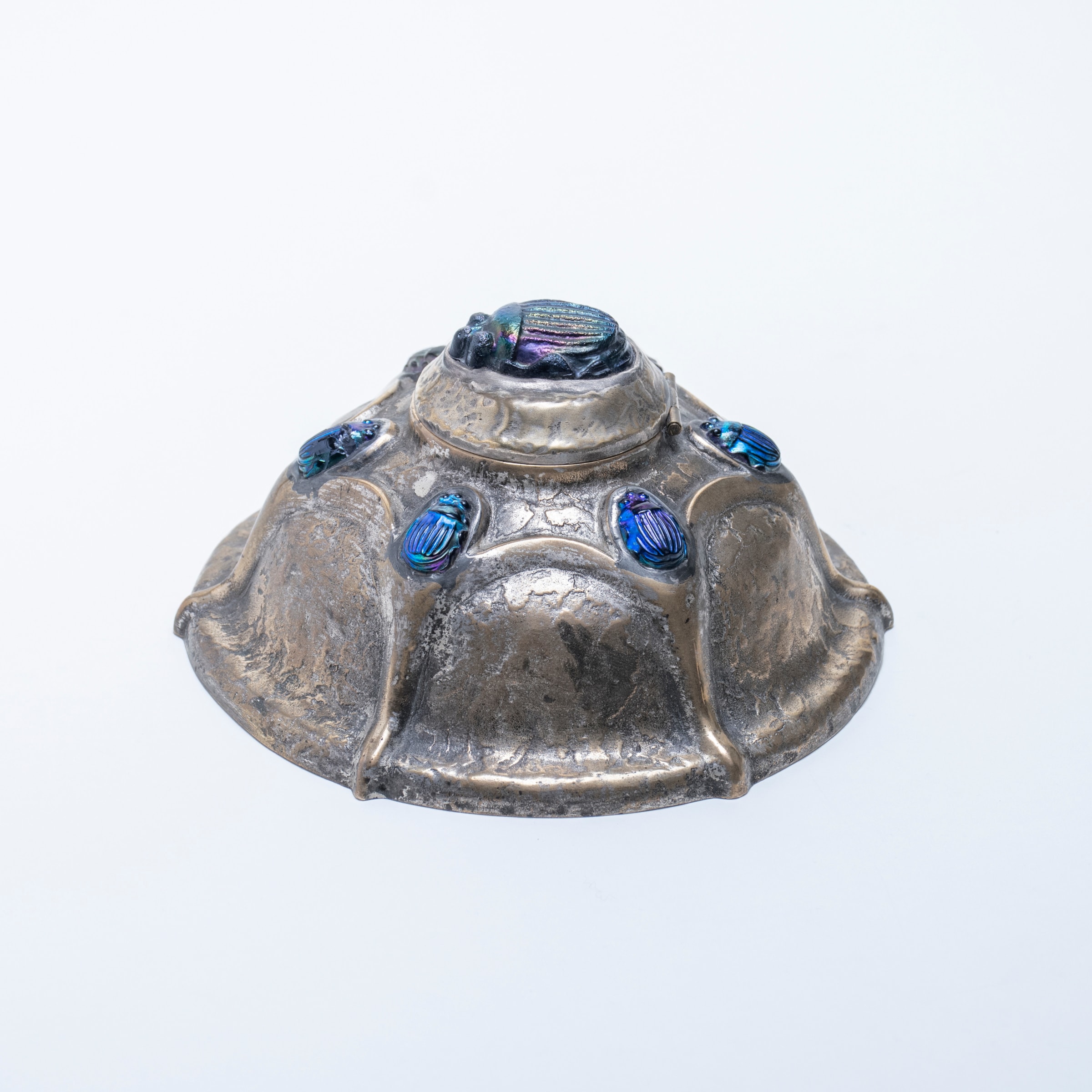 an inkwell by tiffany studios in sterling silver, with irregular surface texture, the hinged top with a large blue iridescent favrile glass pressed scarab tile with rainbow iridescence, with a series of smaller matching tiffany glass scarabs on the domed body of the inkwell.