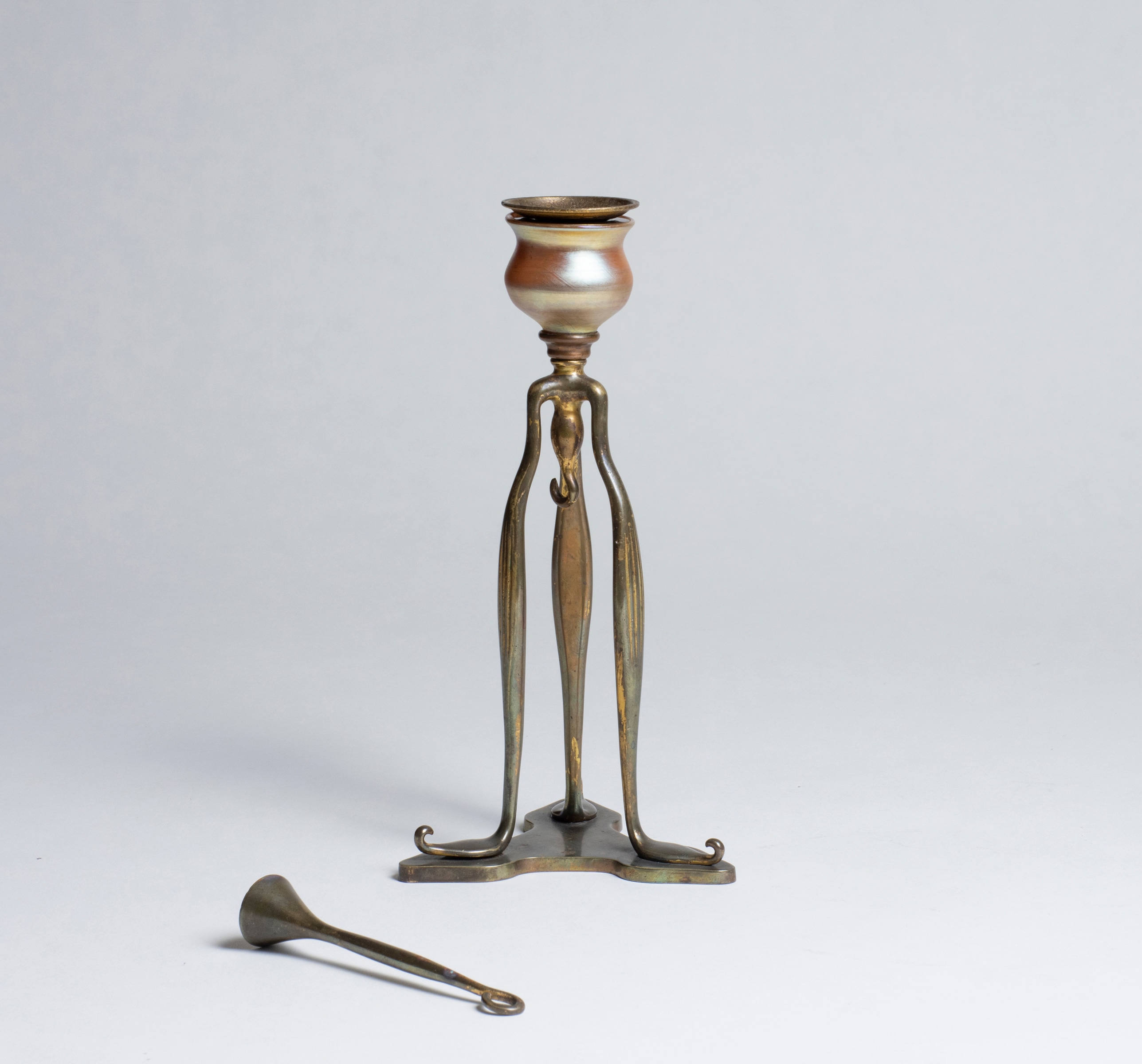 A tiffany studios candlestick, the bronze tripod form with a gold iridescent finial as part of the candle cup; here, showing the matching snuffer which hangs from a hook in the base.