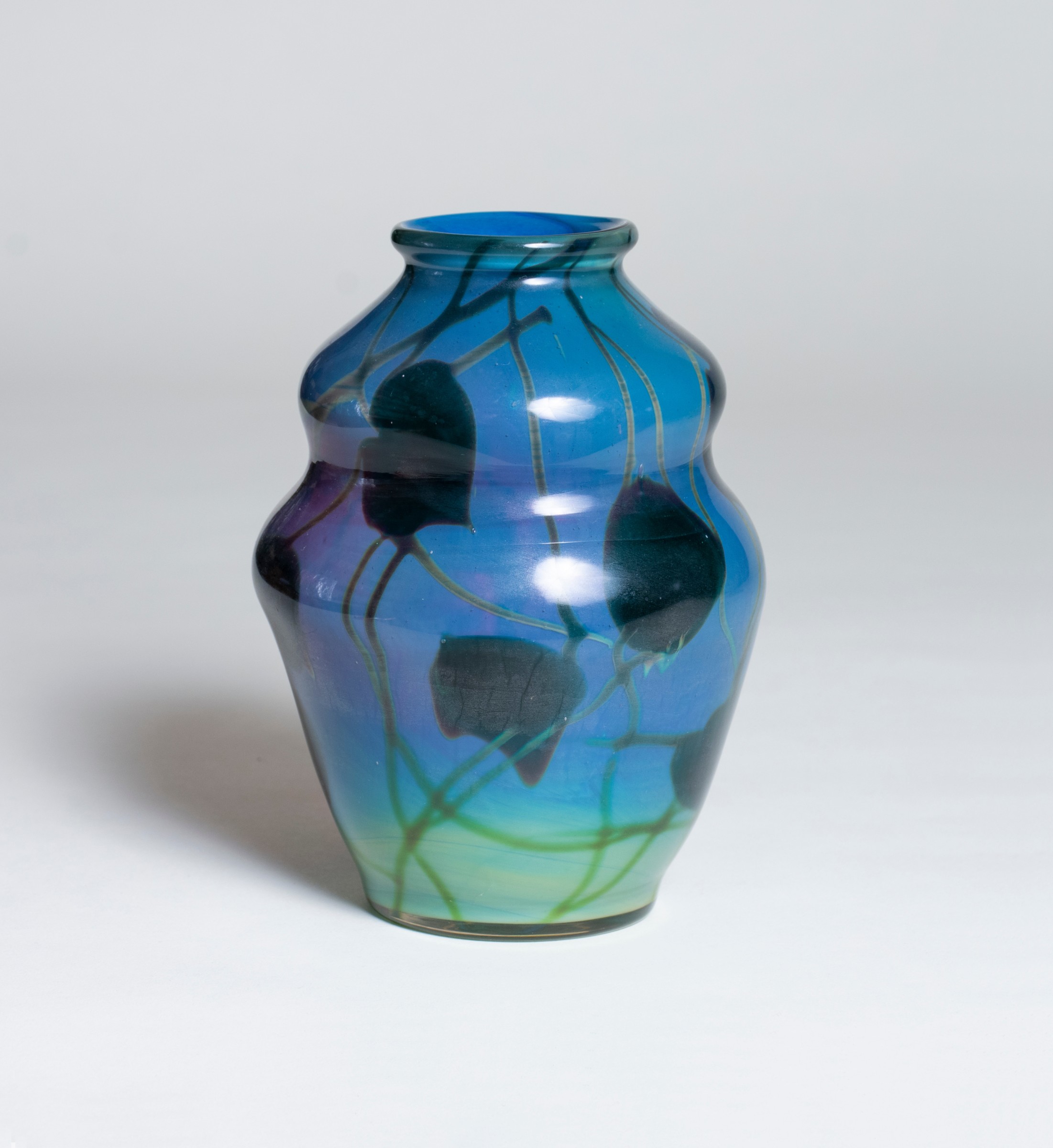 A tiffany favrile glass reactive paperweight style vase, the blue double gourd shaped body shifting in color and opacity as you move around the vase, decorated with all over motif of swirling vines and heart shaped leaves in a dark green glass. The vase appears to be solid and a brighter color blue in reflected light.