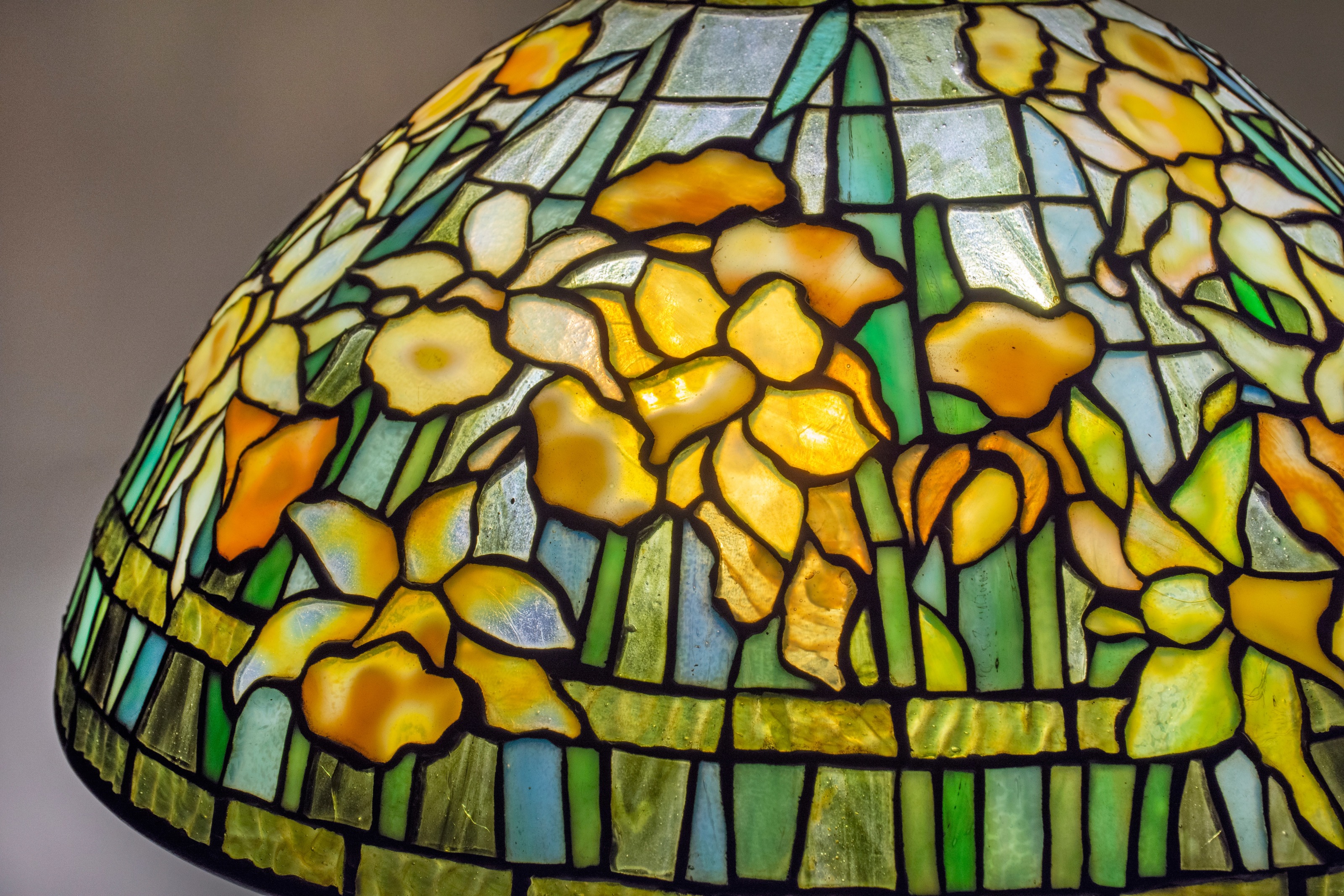 close up of a leaded glass tiffany studios lamp shade showing yellow daffodils in mottled glass, with spiky greenish blue leaves, against a background of translucent glass &quot;sky&quot;