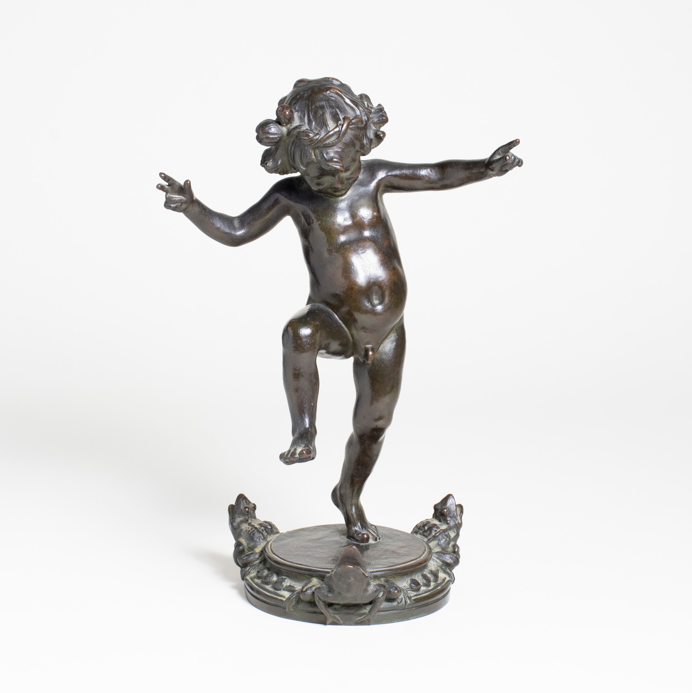 a sculpture of a playful small child frolicking on one foot, the other leg raised, the arms outstretched in a dance like gesture, smiling and laughing. the circular base of the statue features three small frogs and functions as a fountain
