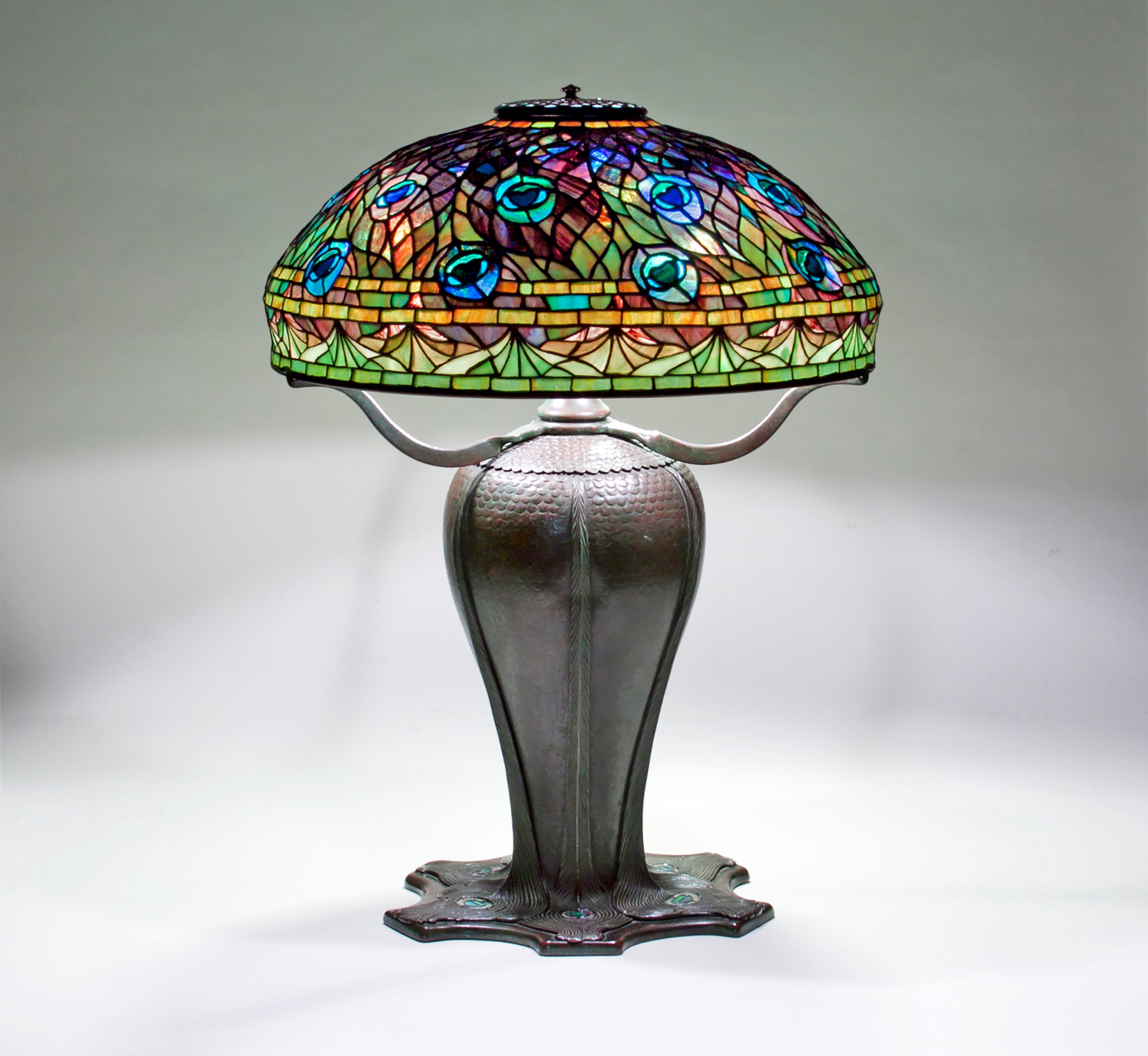 a tiffany table lamp, the dome shaped shade in leaded glass design depicting stylized swirling peacock feathers with bright blue eyes, the rest in shades of dark purple near the top shifting through orange and gold to green near the lower edge