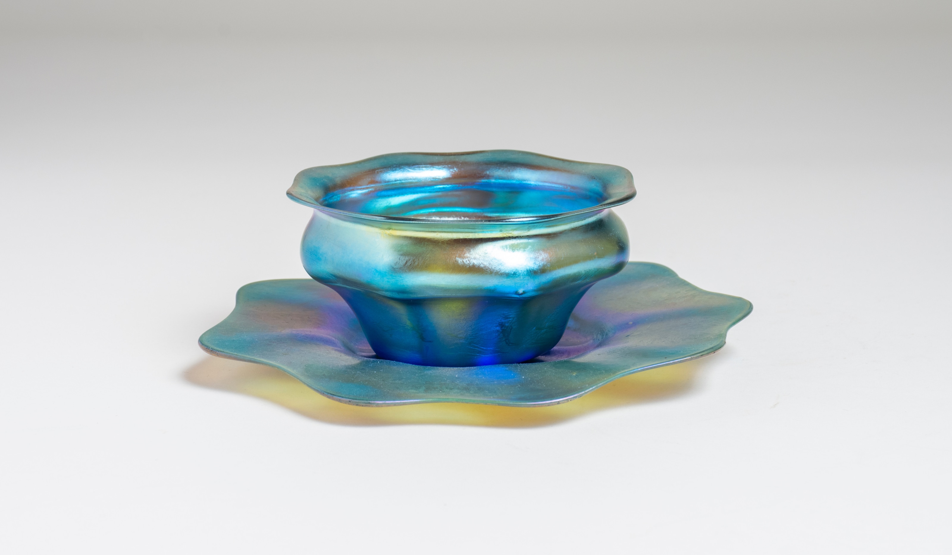 a blue iridescent favrile glass finger bowl and underplate by tiffany studios, the underplate in extremely thin ribbed glass with a waving edge, the small ribbed bowl in matching blue glass fitting into the plate at the center.
