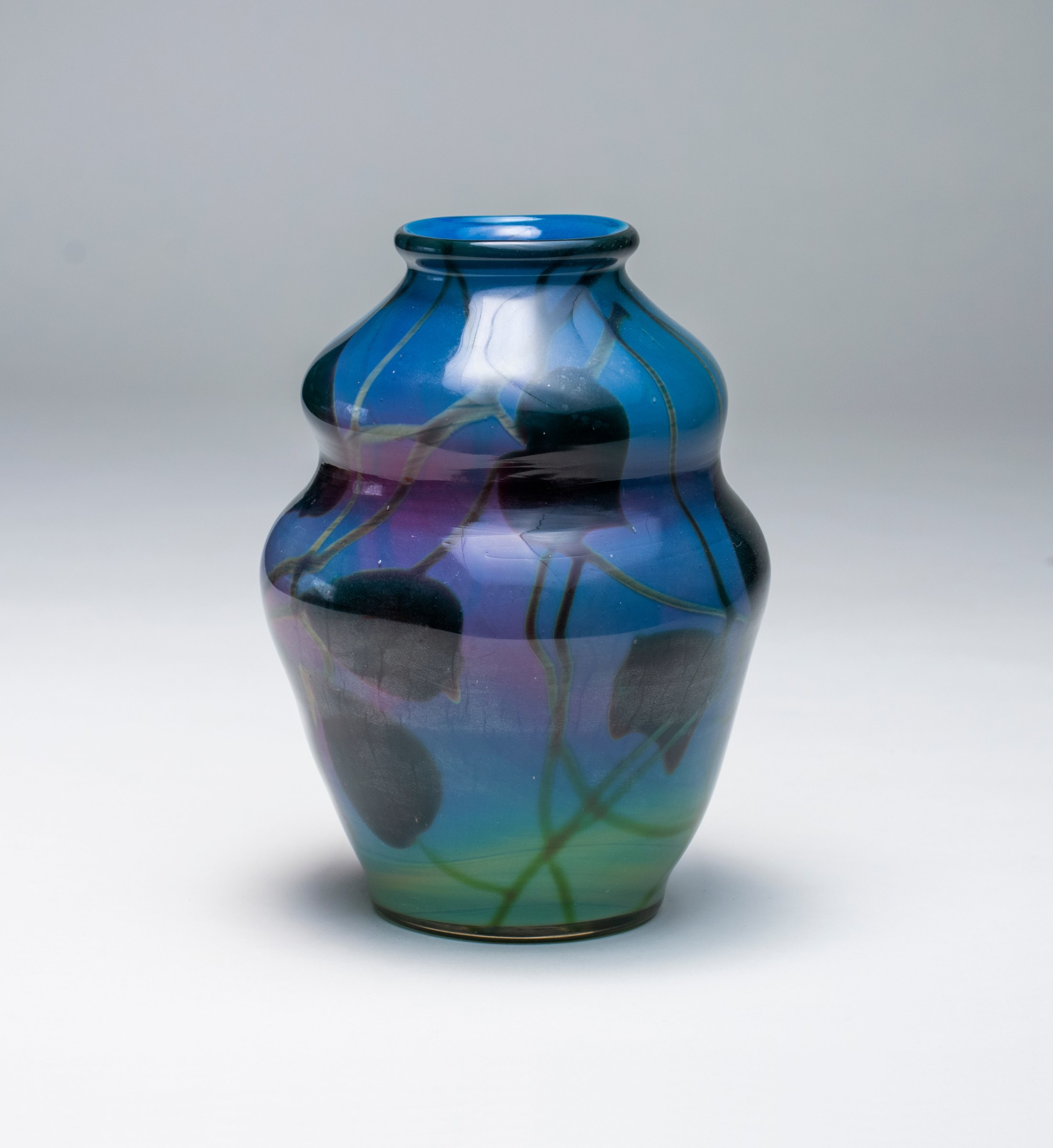 A tiffany favrile glass reactive paperweight style vase, the blue double gourd shaped body shifting in color and opacity as you move around the vase, decorated with all over motif of swirling vines and heart shaped leaves in a dark green glass. The vase appears to glow purple in transmitted light.