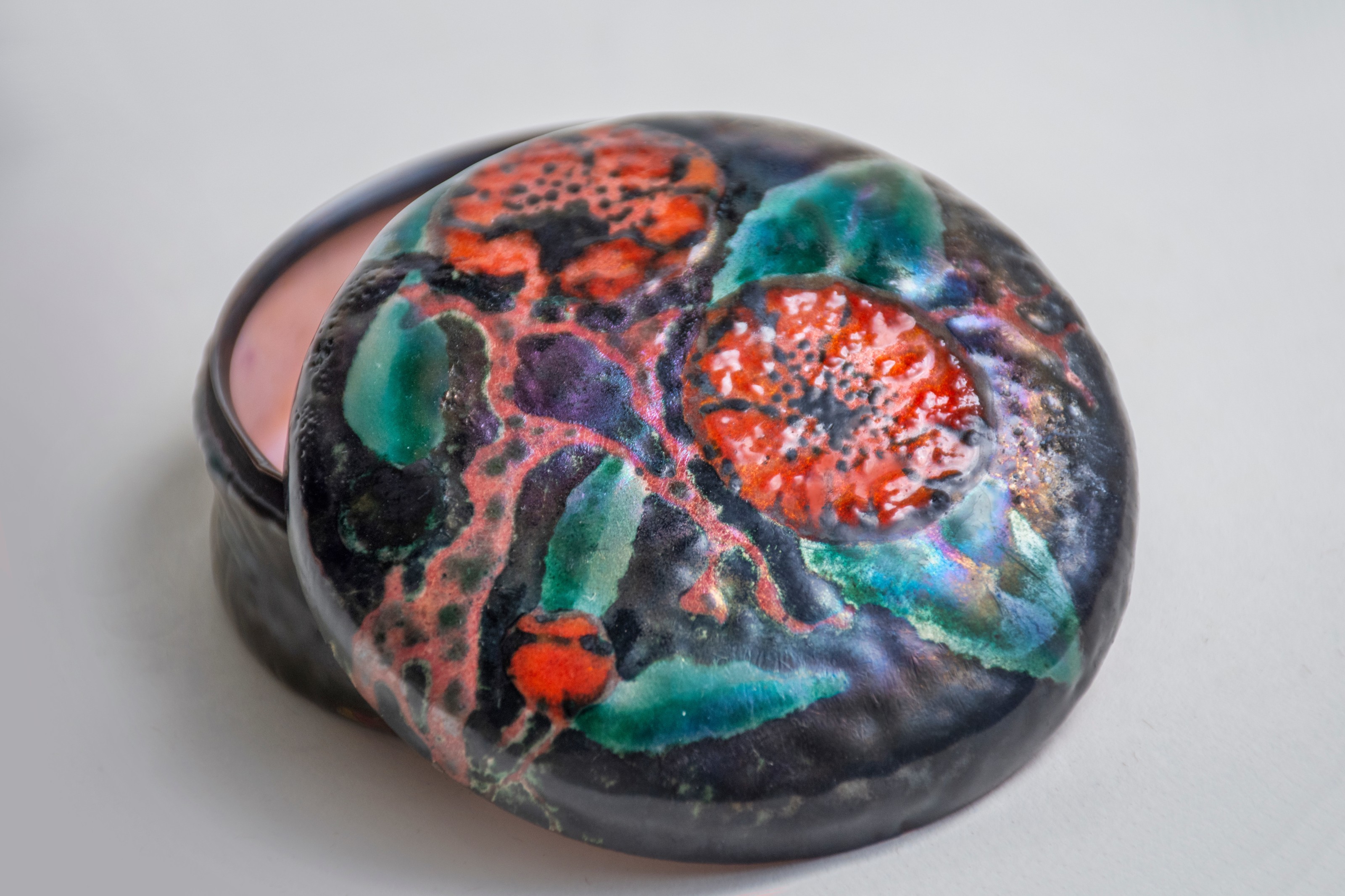 A close up showing the lid of the enameled box showing the iridescent finish of the enamels used in the poppies and their leaves, with raised patterning on the flower centers.