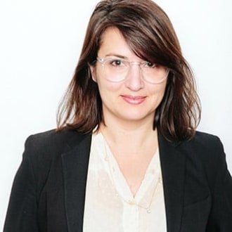 The torso of a woman with medium fair skin, dark hair and brown eyes looks directly at us on a white background. She is wearing clear geometric glasses and a white blouse with a black blazer. 