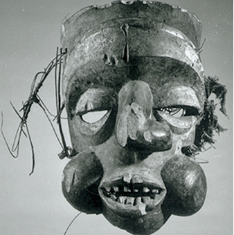 Mask, Ibibio. Mask made of wood, stain, reed, and fabric, replicating a human profile, with eyes carved out beneath the eyelids. The mouth is open, bearing teeth, and its bulbous cheeks and twisted nose greatly protrude from the mask. 