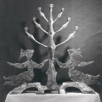 Black and white image of a bronze menorah. On each side of the menorah is a female figure with one arm reaching for the stem of the menorah and the other outstretched, while legs are outstretched on the base. 