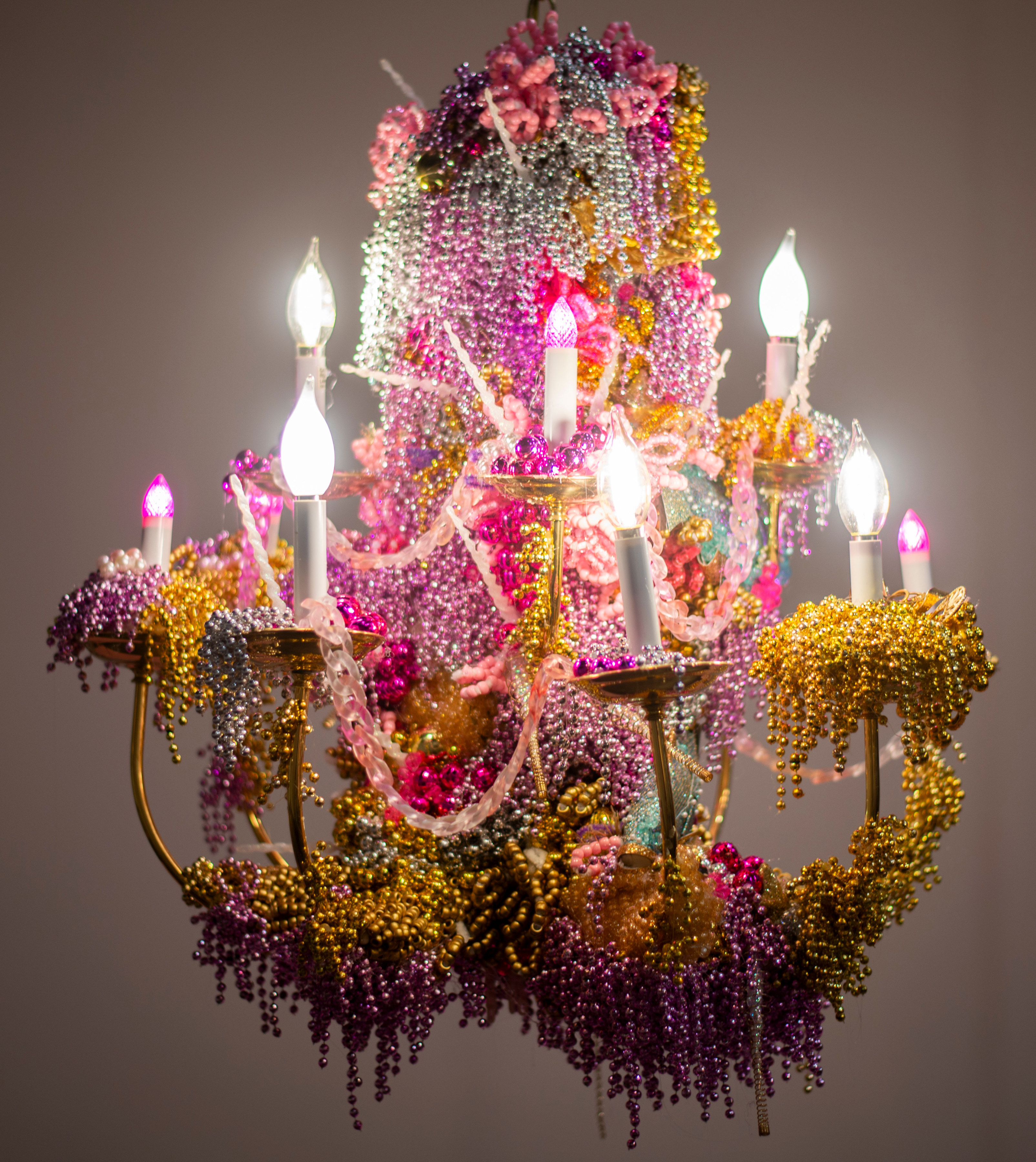 A chandelier made by Jenny Day coated with Mardi Gras beads and sequins, emitting a warm glow from its electric candles.