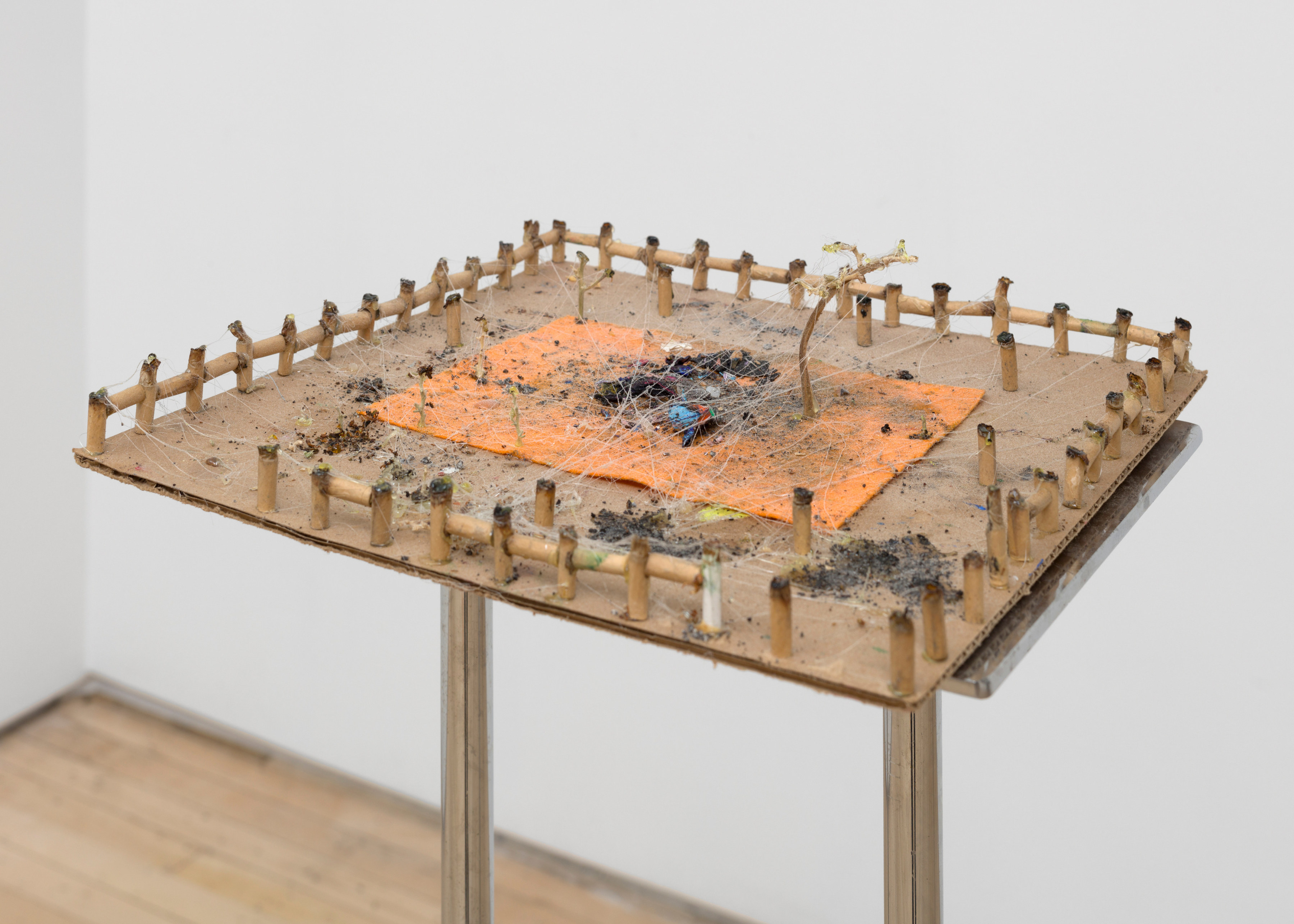 A sculpture by JPW3 of the ends of marijuana joints linked together making a miniature series of fences on a cardboard panel atop a shining metal platform on a white pedestal.