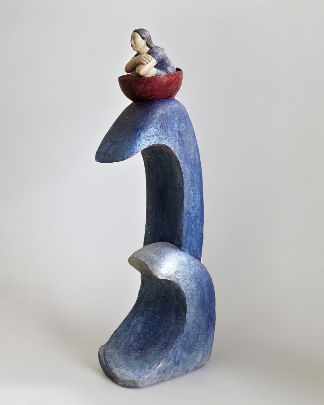 A sculpture of a woman with her arms crossed in a burgundy bowl atop large blue waves. 