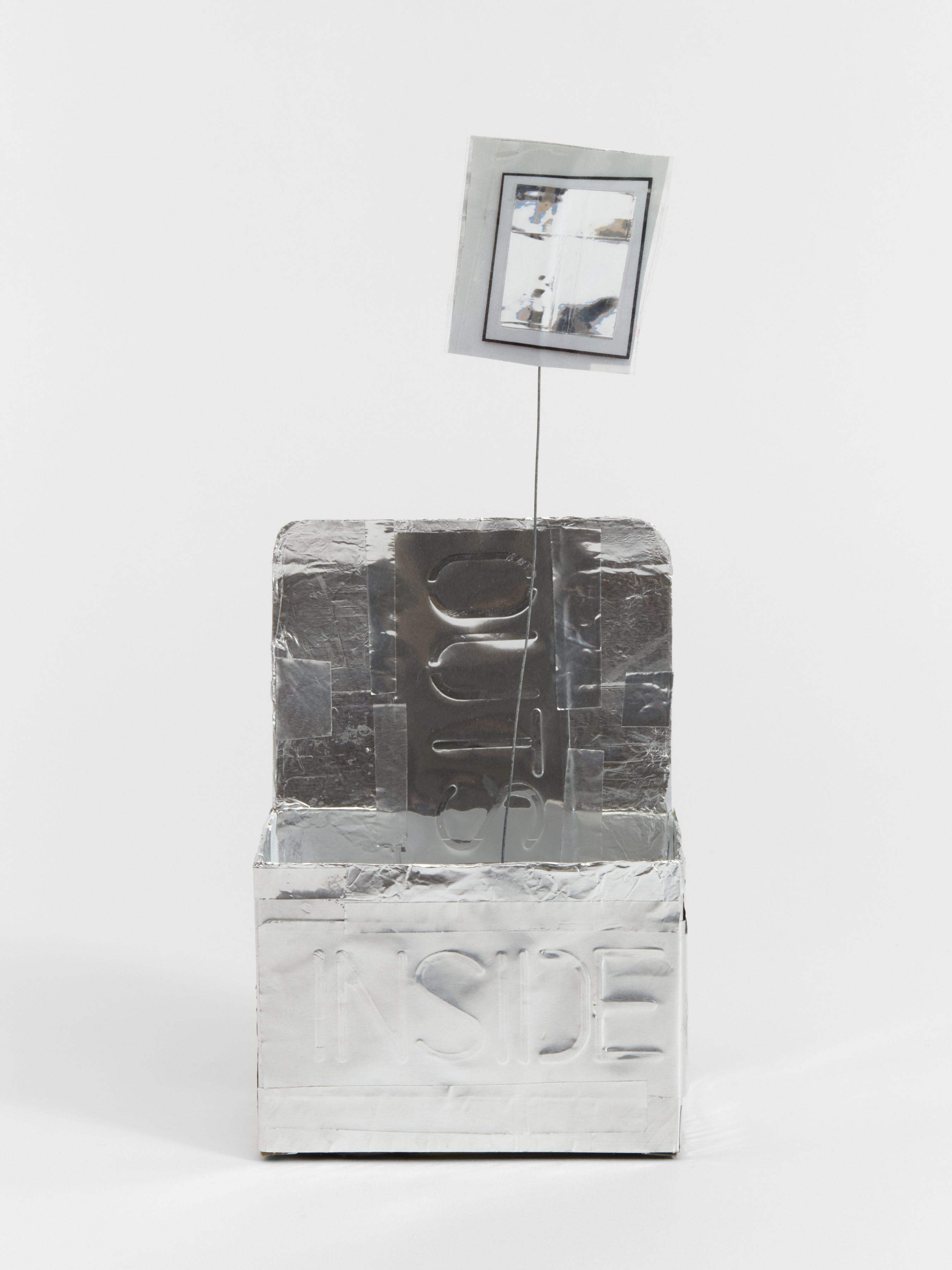 A box like sculpture with a mirror on a wire emerging from the cavity coated in a reflective material with the words "inside" and "out" embossed on the surface. 