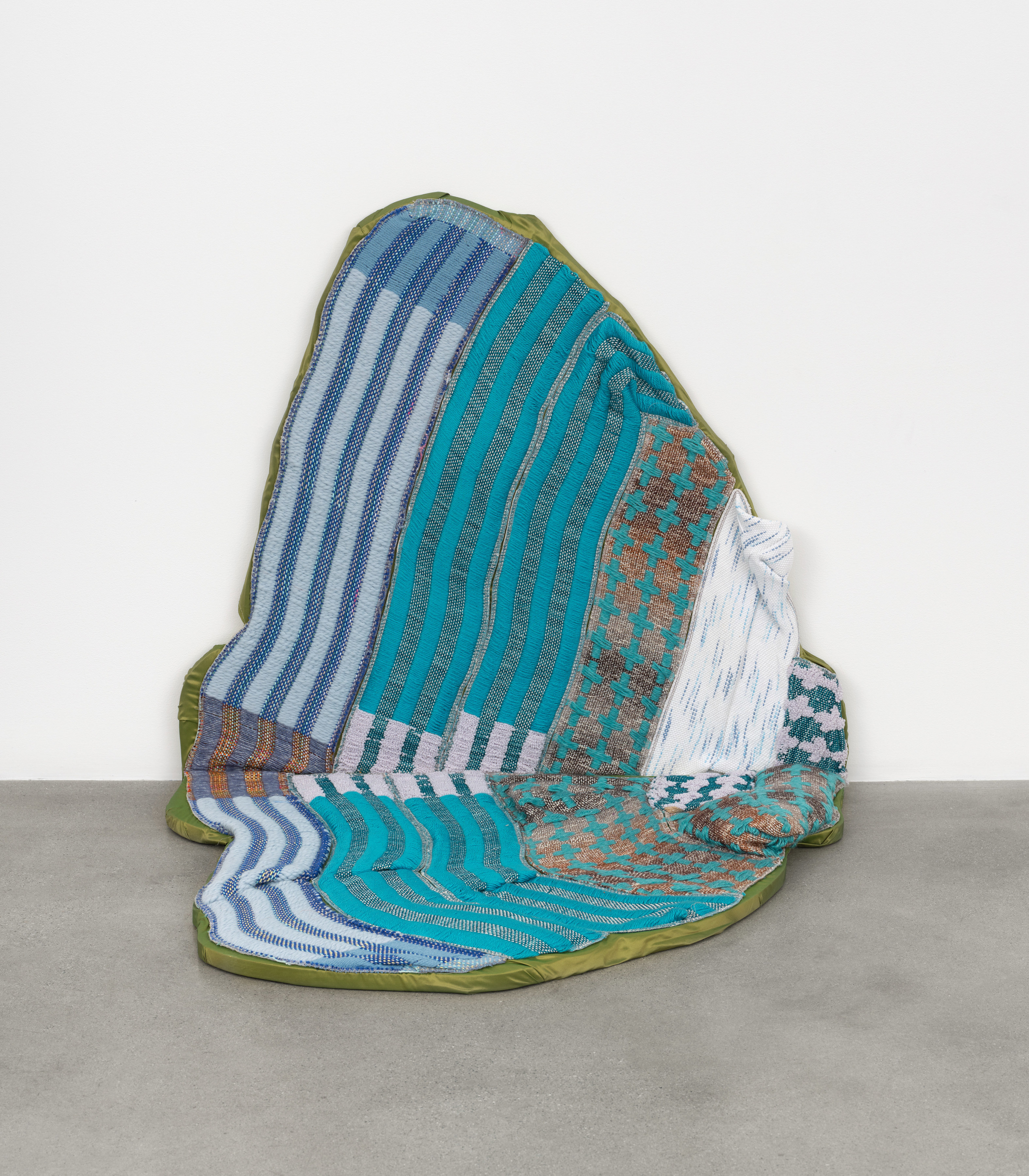 A soft textile sculpture of an oblong oval shape in brown and blue fabric resting between the floor and the gallery wall. 