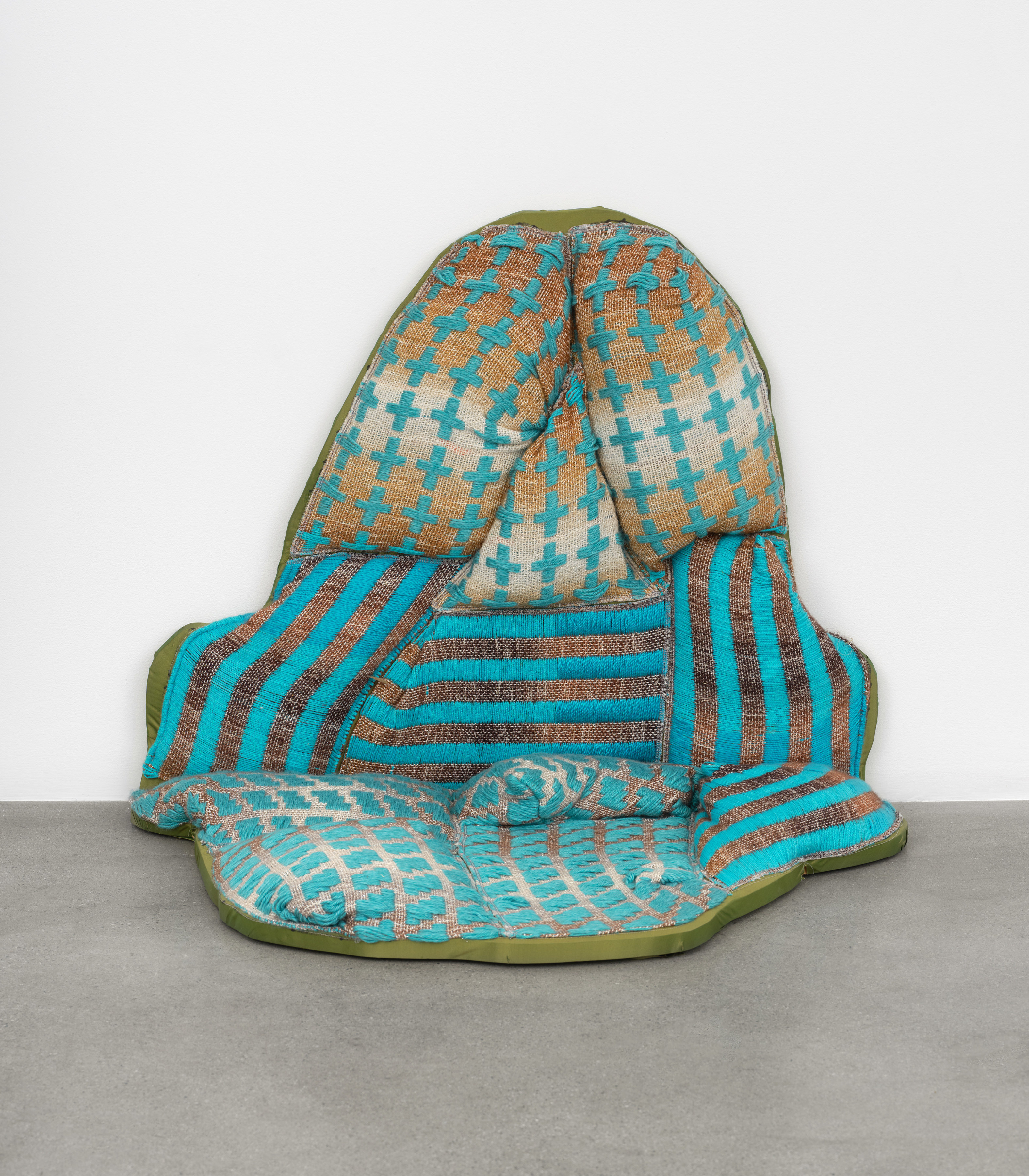 A soft textile sculpture of an oblong oval shape in brown and blue fabric resting between the floor and the gallery wall. 