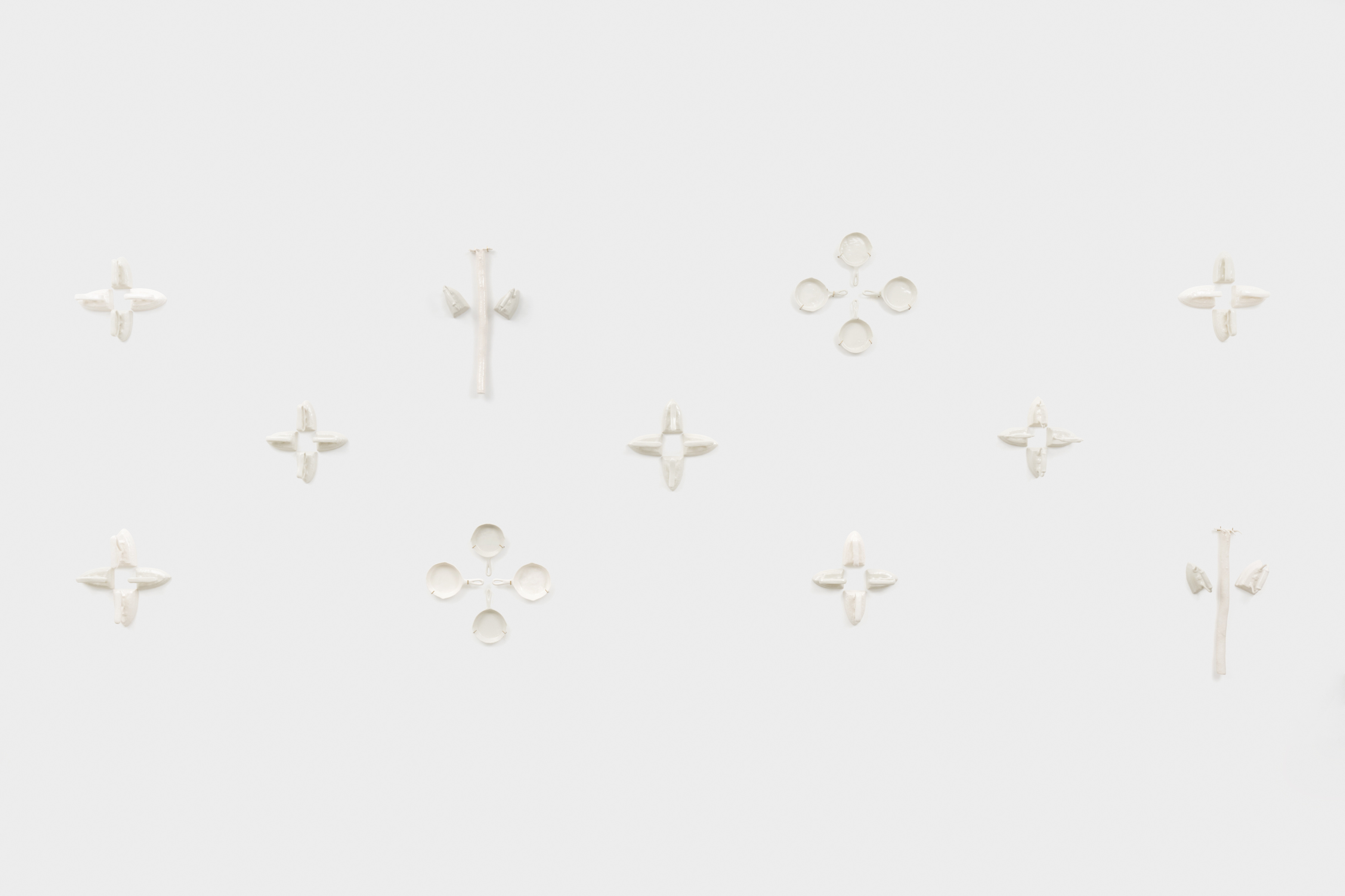 A multi piece wall mounted sculpture with ceramic replicas of irons, cast iron pans and rakes hung in groups of three and four along the wall making a star like pattern.