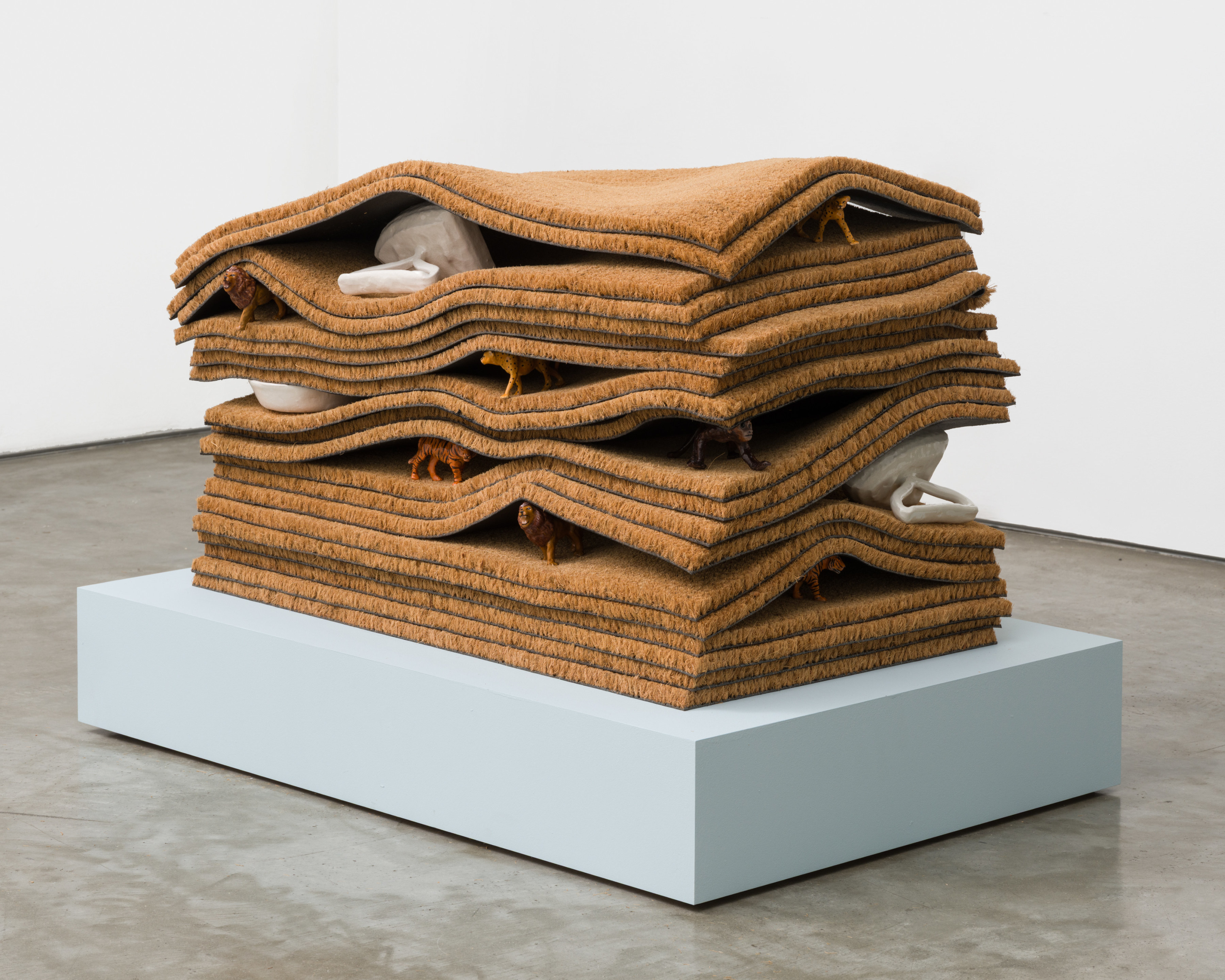 A sculpture with 20 layered doormats with ceramic animal figurines and ceramic irons peaking out from between the layers.