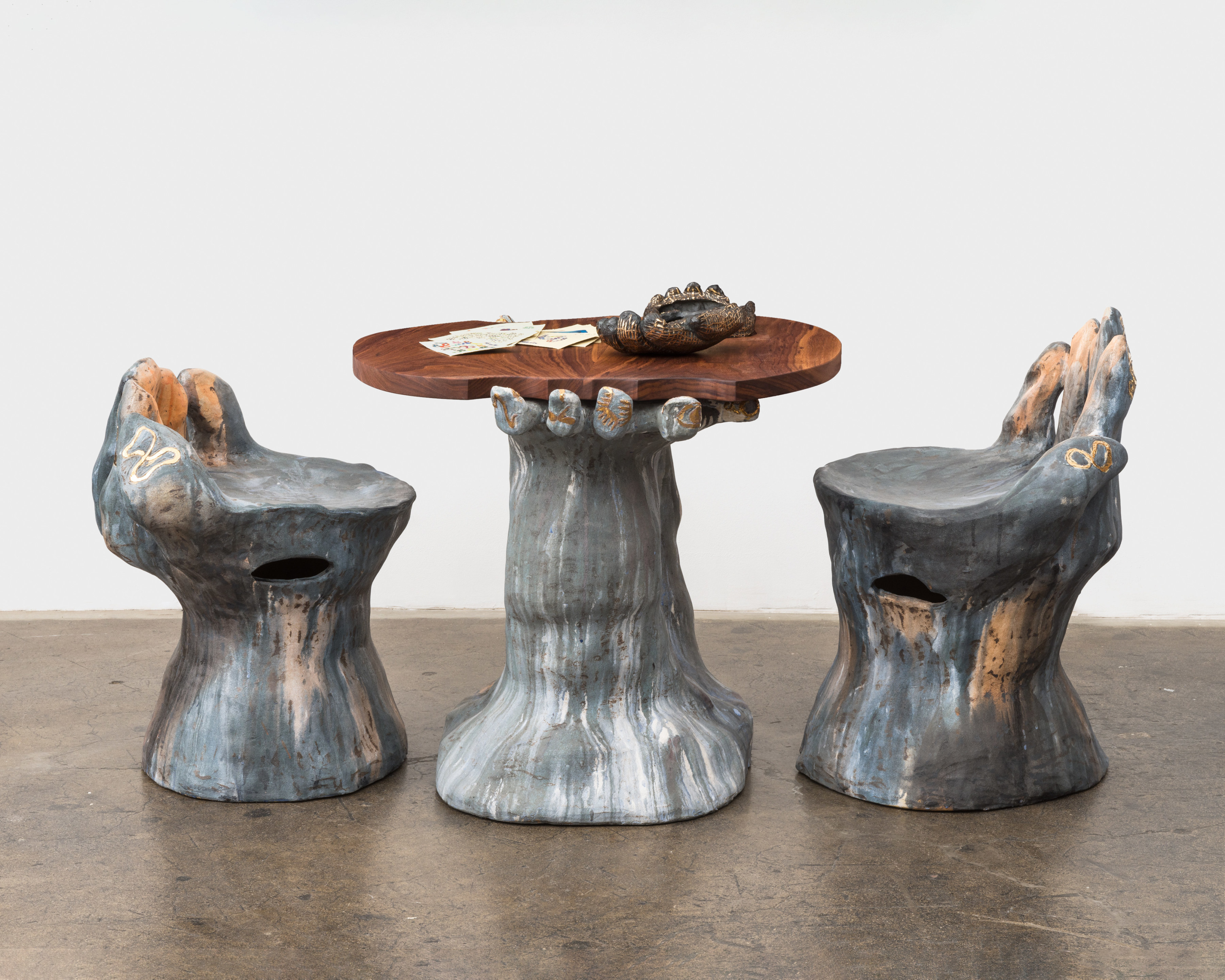 A three piece ceramic sculpture which includes two large grey and pink glazed hand shaped chairs and a walnut table top held by two ceramic hands.