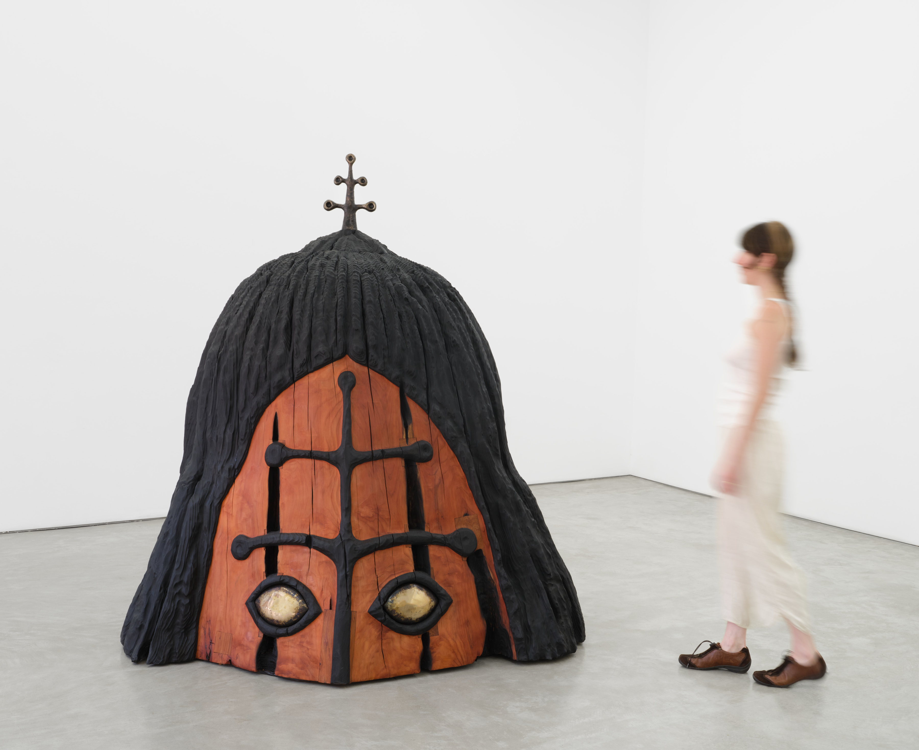 A redwood sculpture, a large top portion of a face. Black symmetrical design on the center, with aluminum eyes. And a metal antenna figure peeking out from the top of the head.