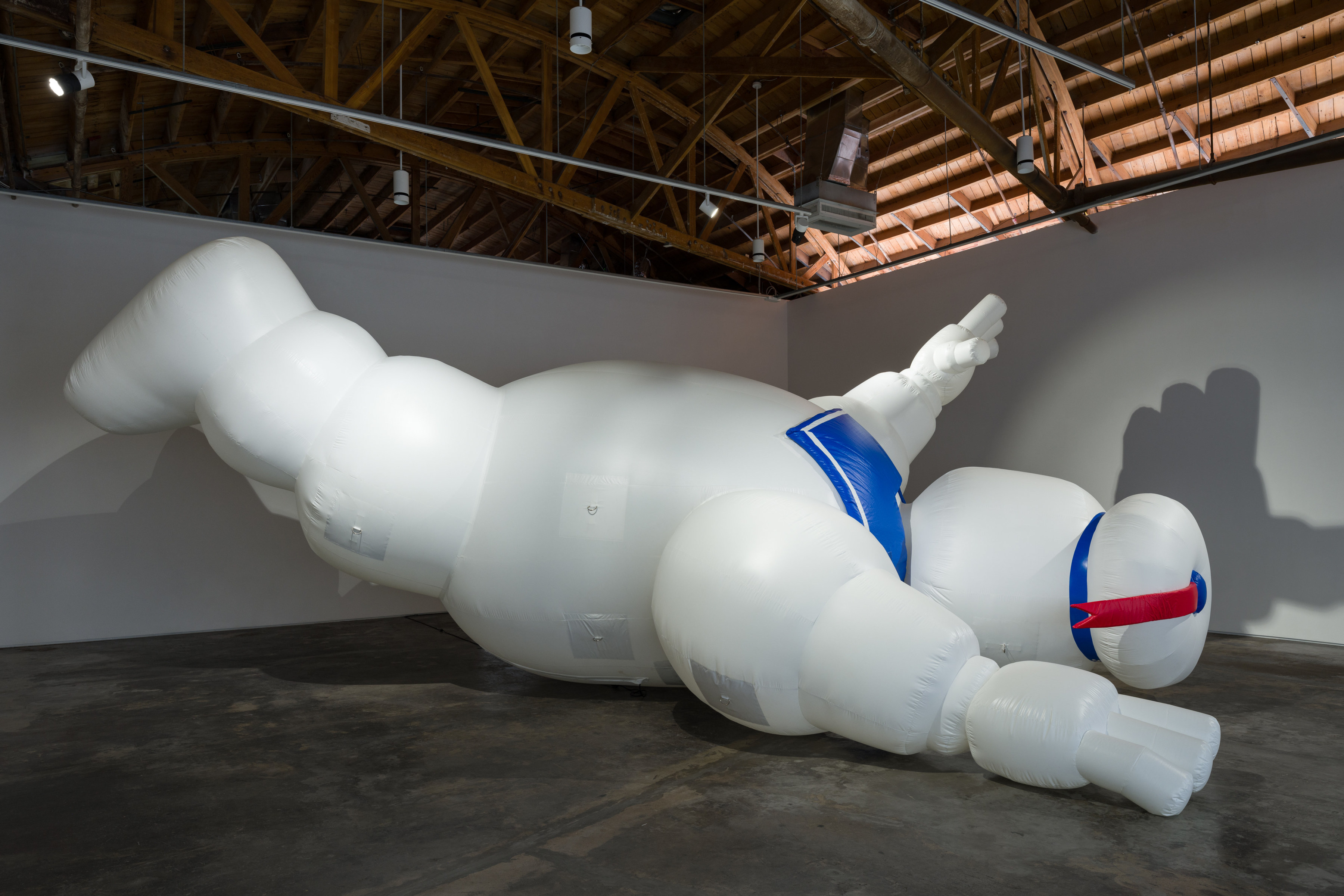 A 30 foot long and 21 foot high inflatable sculpture of the "StayPuft" character from the Ghostbusters movie franchise that who is white and bulbous with a sailor's collar and red tailed cap face down, feet in the air and left arm raised.