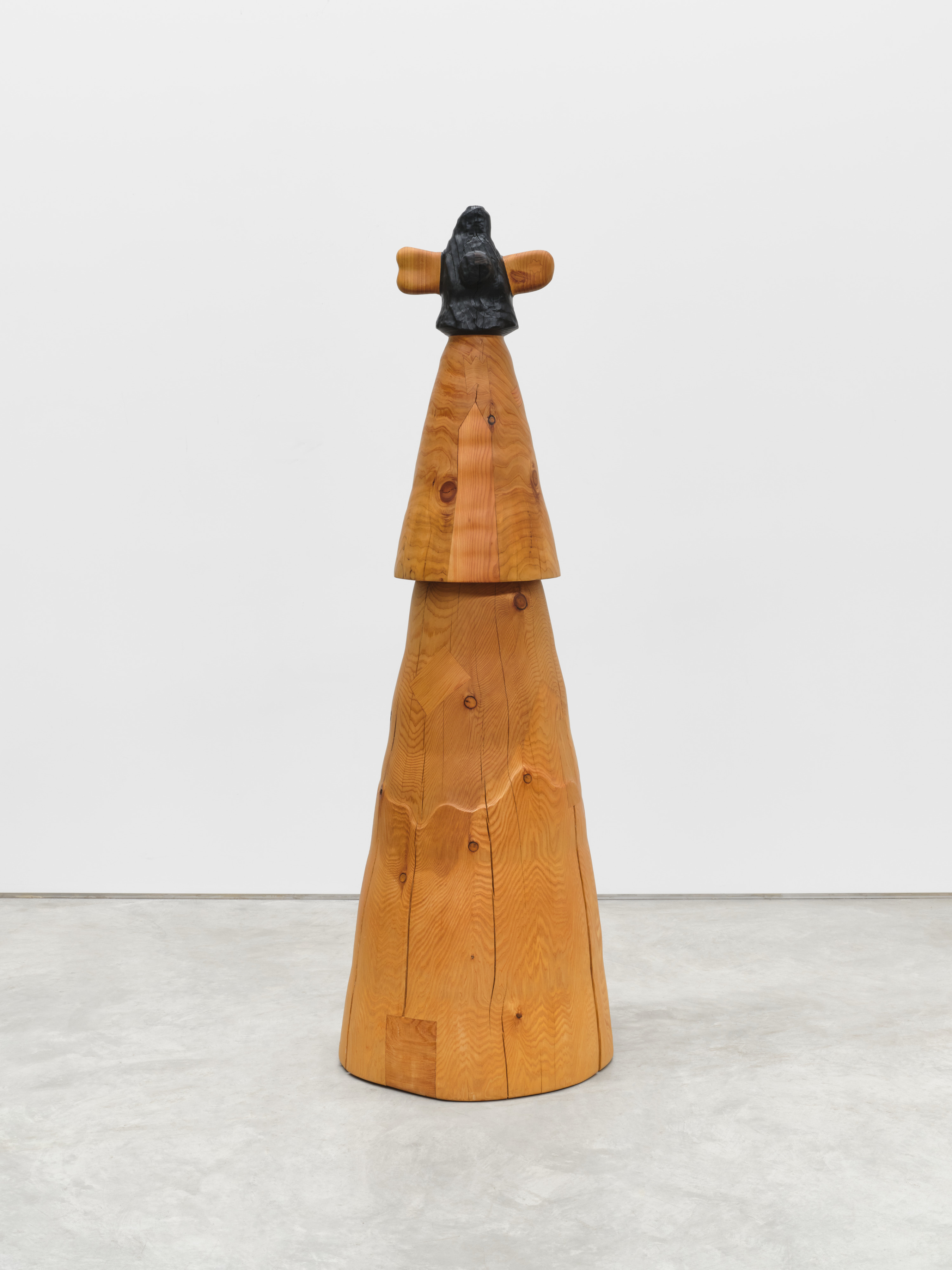 A three tiered carved wooden sculpture at human scale with a large cone shaped base, a smaller cone shaped center and a blackened cone shaped top with two oblong protrusions in a natural wood grain.