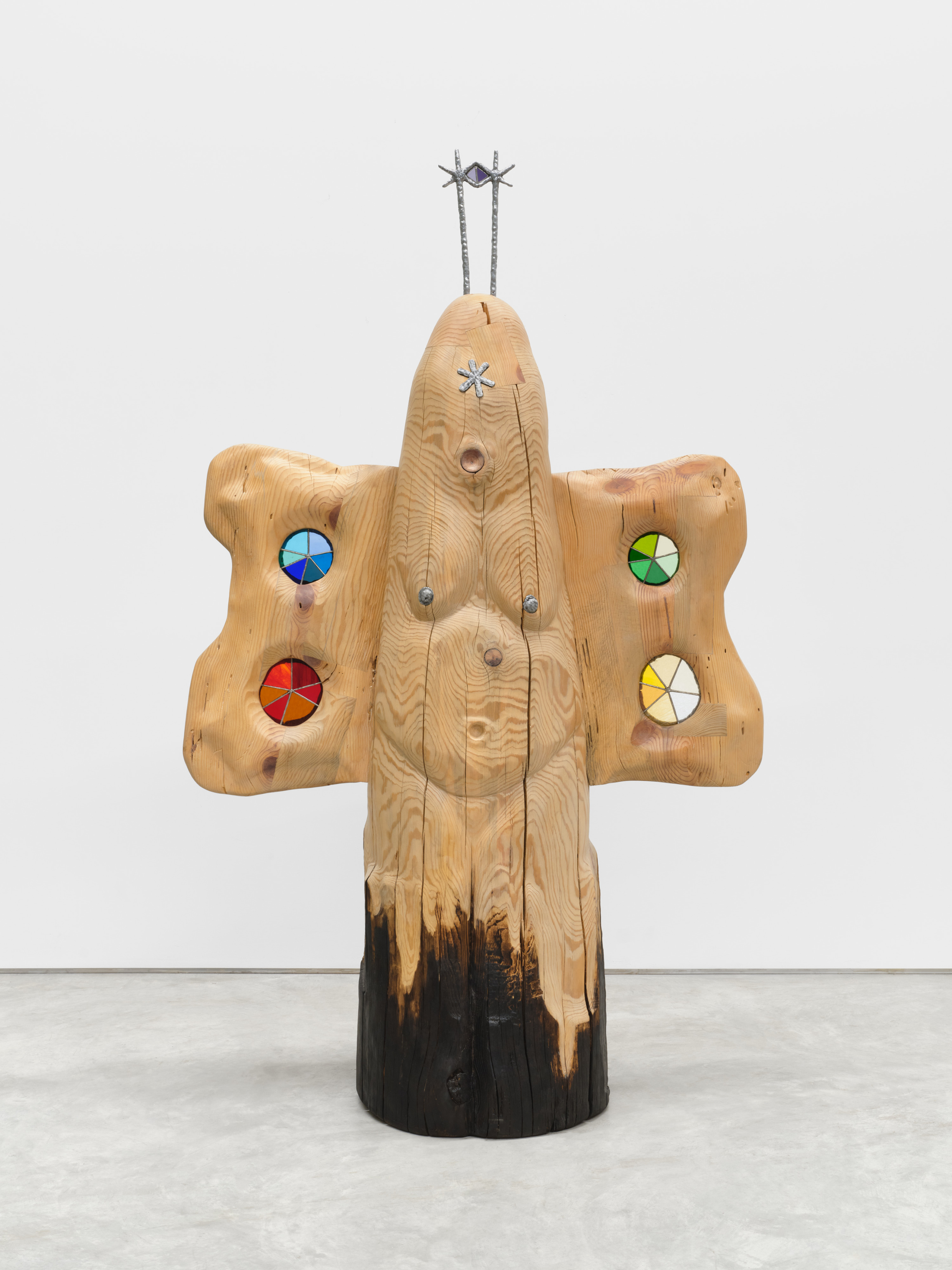 A carved anthropomorphic wooden sculpture at human scale with wings that have stained glass windows and two metal antennae. 