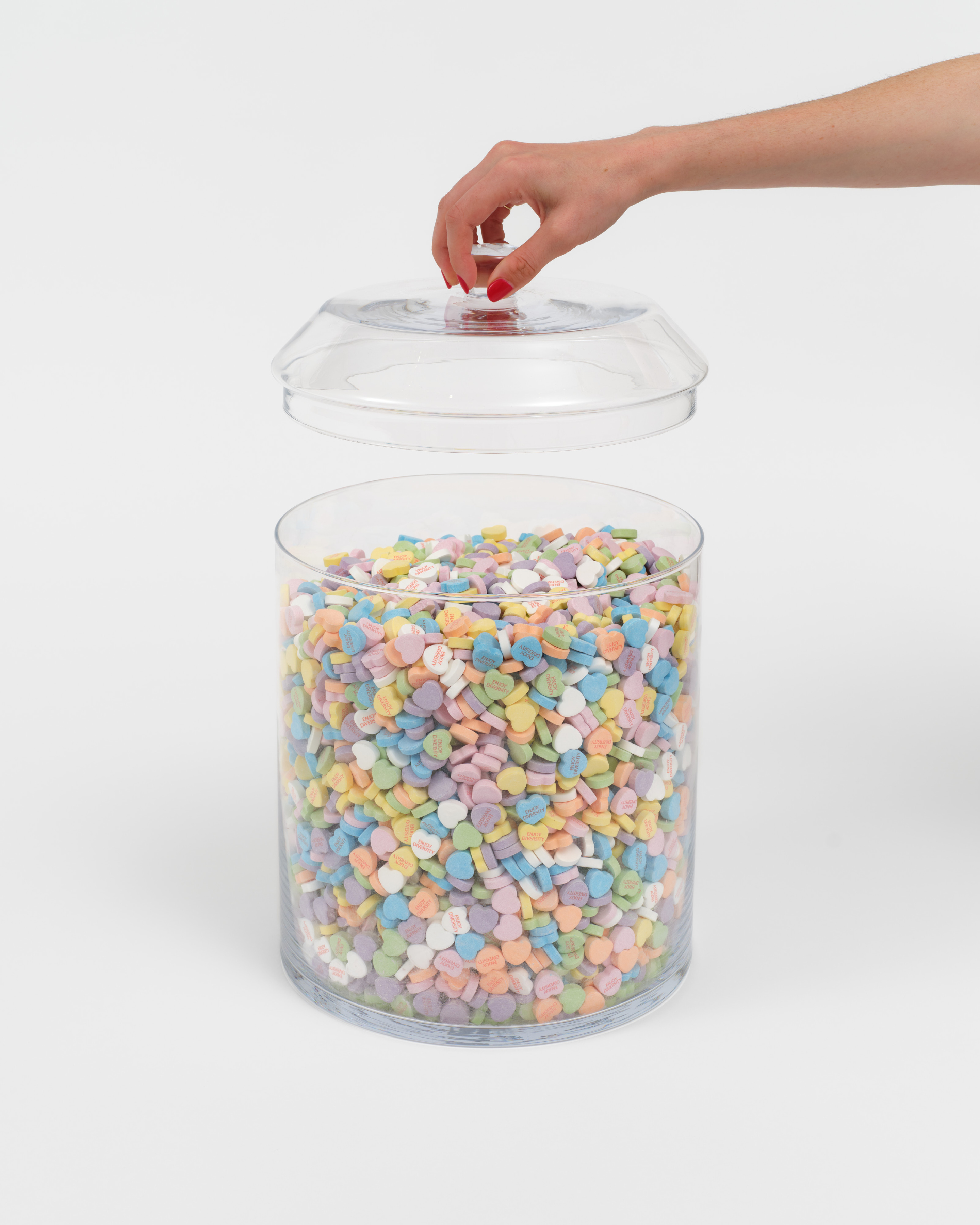 A hand lifts the lid of a glass jar filled with heart shaped candies that read "Enjoy Diversity"