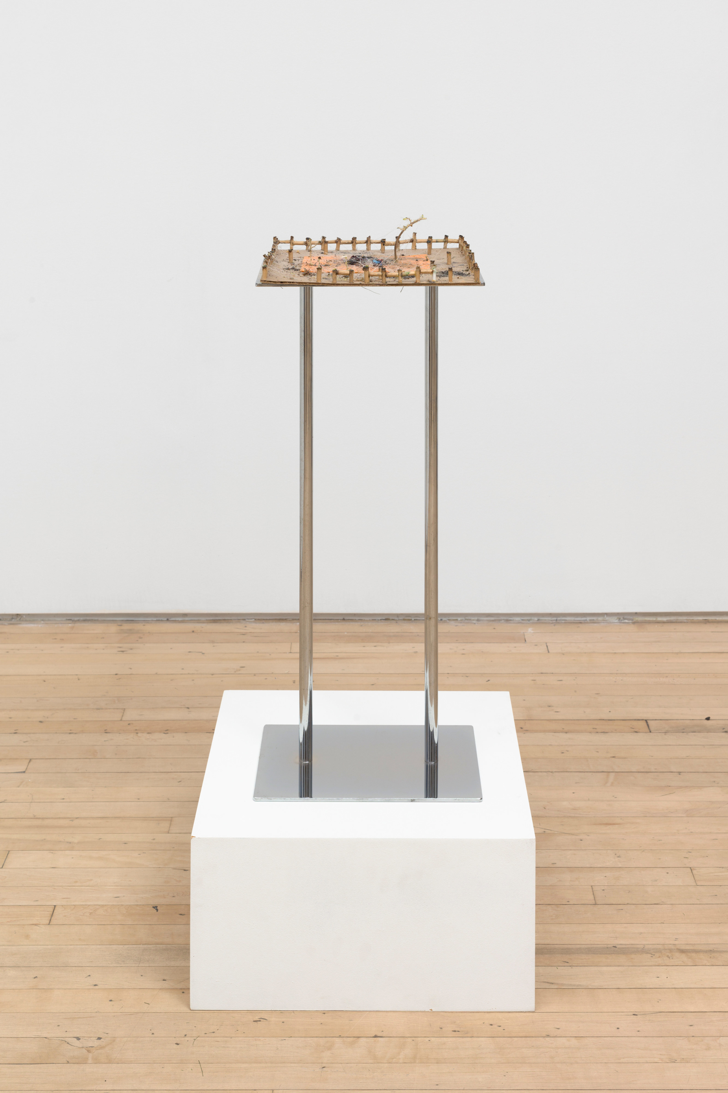 A sculpture by JPW3 of the ends of marijuana joints linked together making a miniature series of fences on a cardboard panel atop a shining metal platform on a white pedestal.