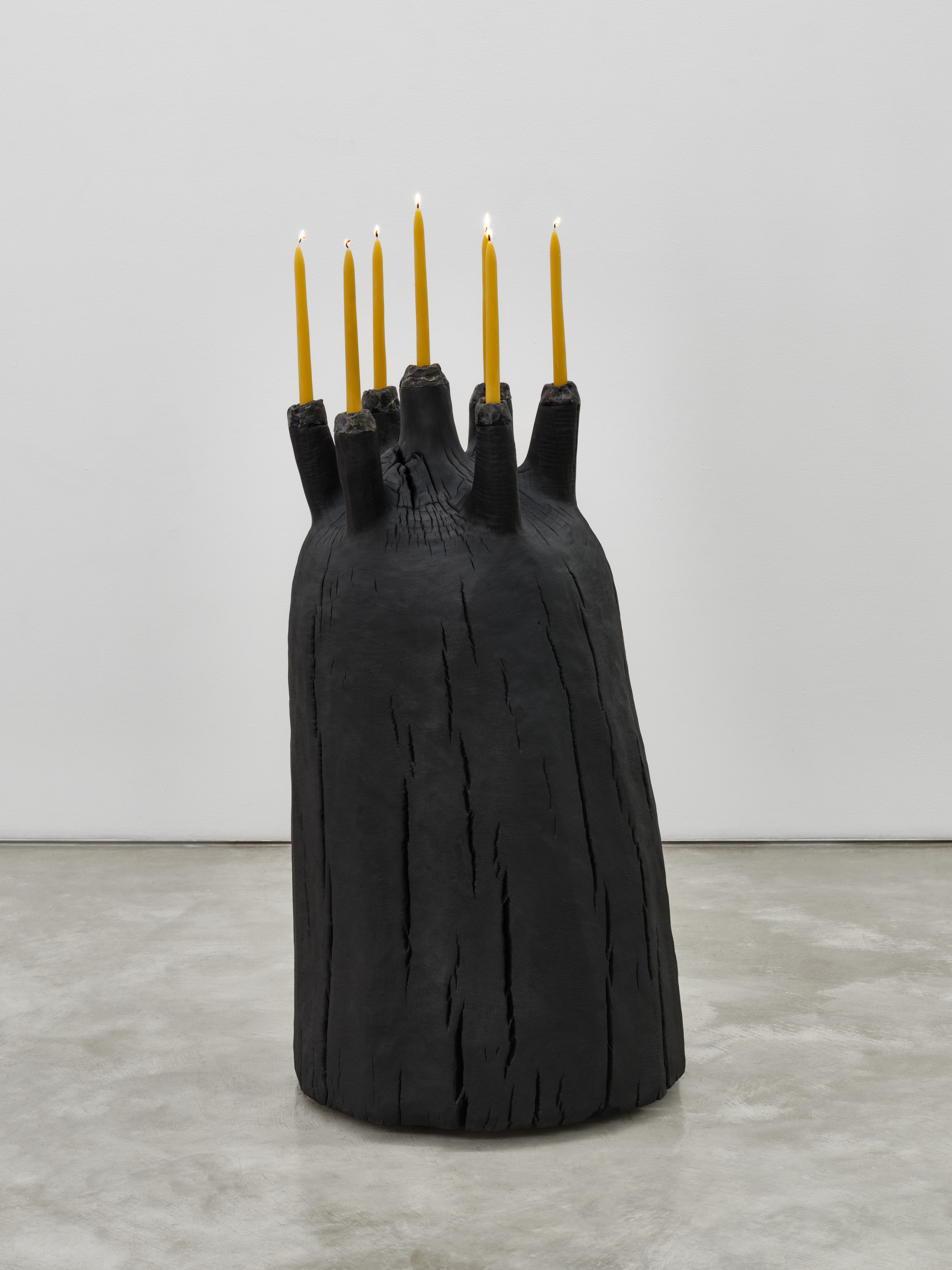 A black mound shaped wooden sculpture with seven long taper candles protruding from the top.