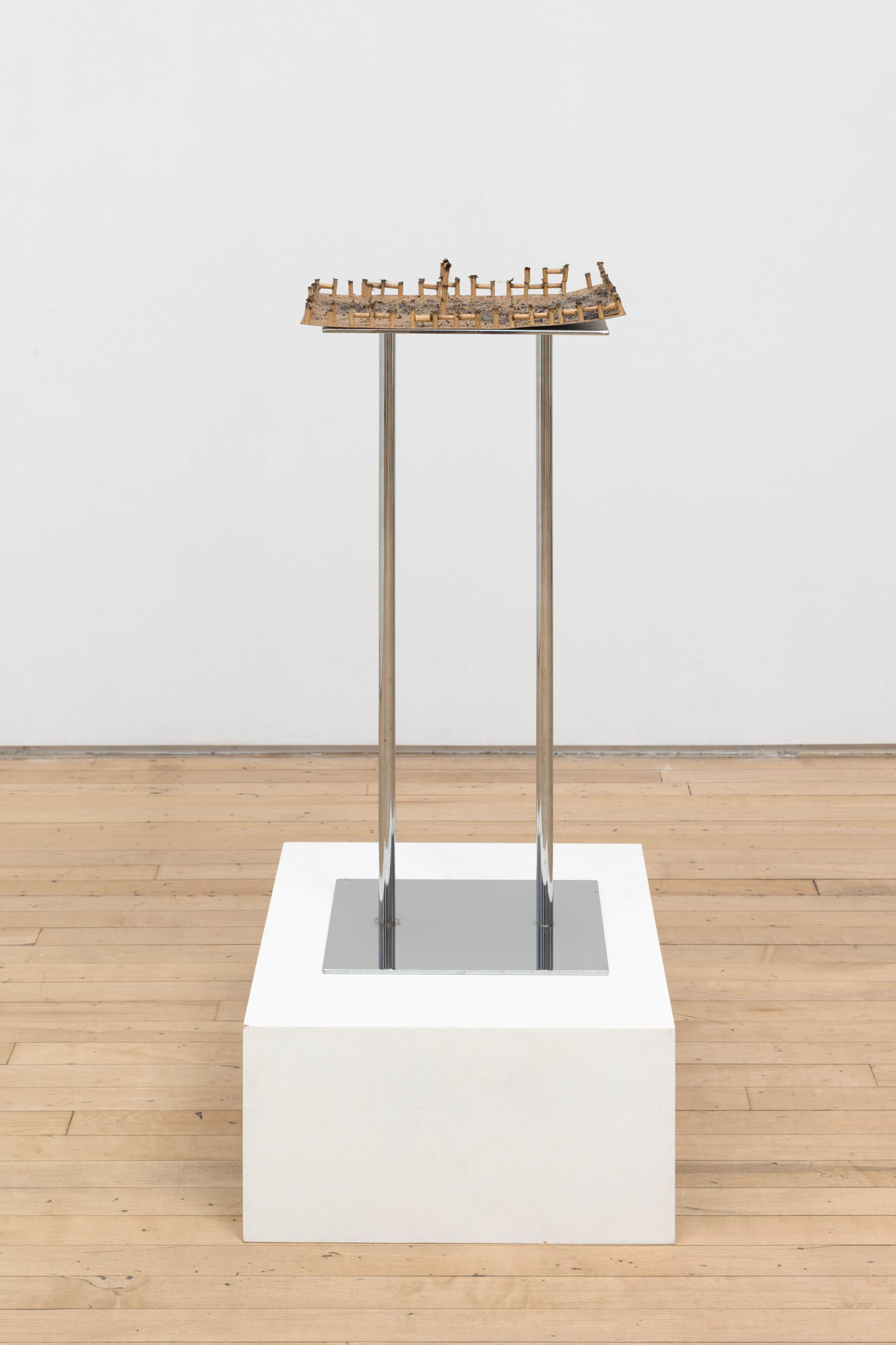 A sculpture by JPW3 of the ends of marijuana joints linked together making a miniature series of fences on a cardboard panel atop a shining metal platform on a white pedestal.