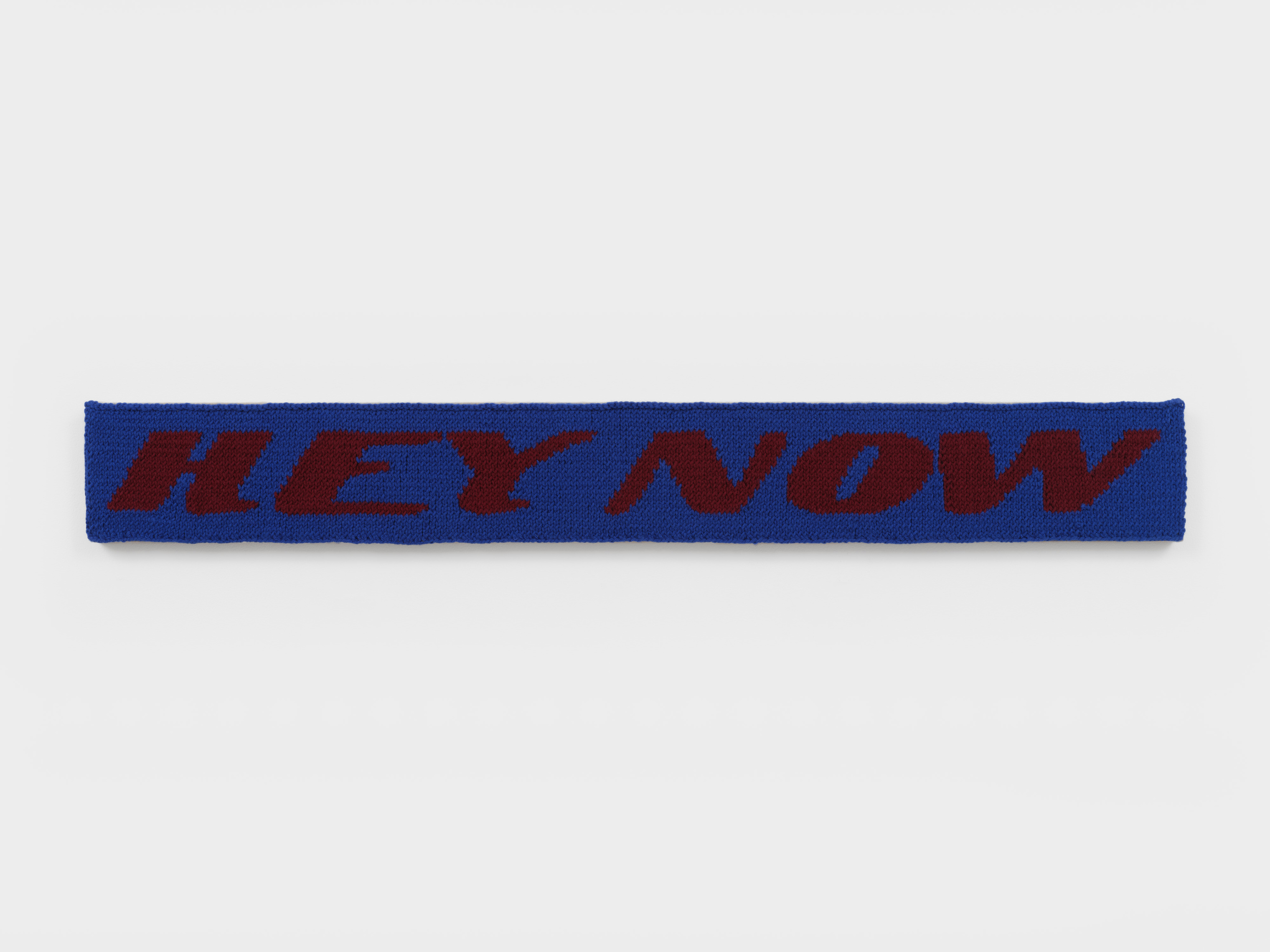 A knit work rendered in blue and burgundy yarn that reads "Hey Now" in a long thin rectangle sewn into linen.