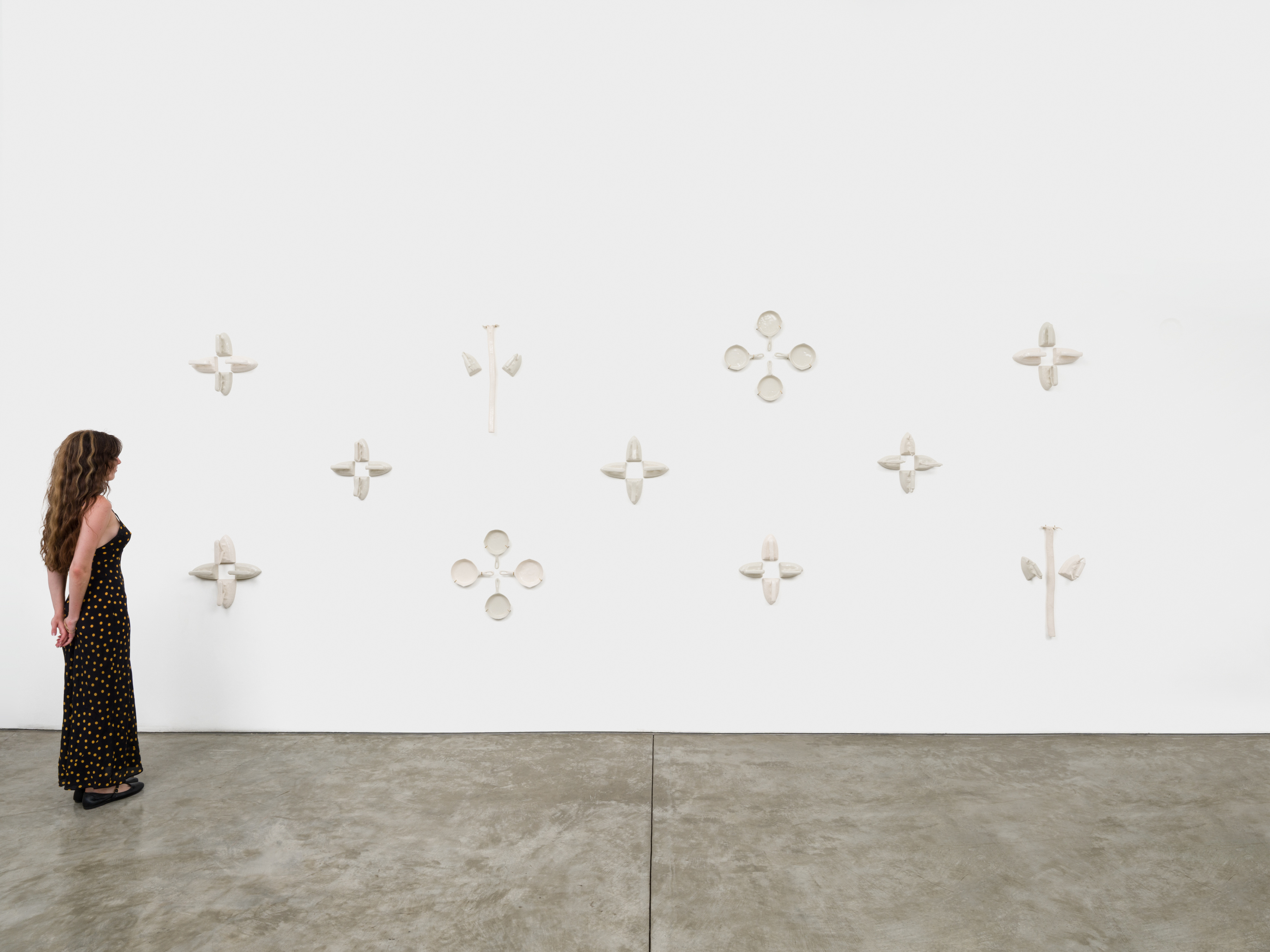 A multi piece wall mounted sculpture with ceramic replicas of irons, cast iron pans and rakes hung in groups of three and four along the wall making a star like pattern.