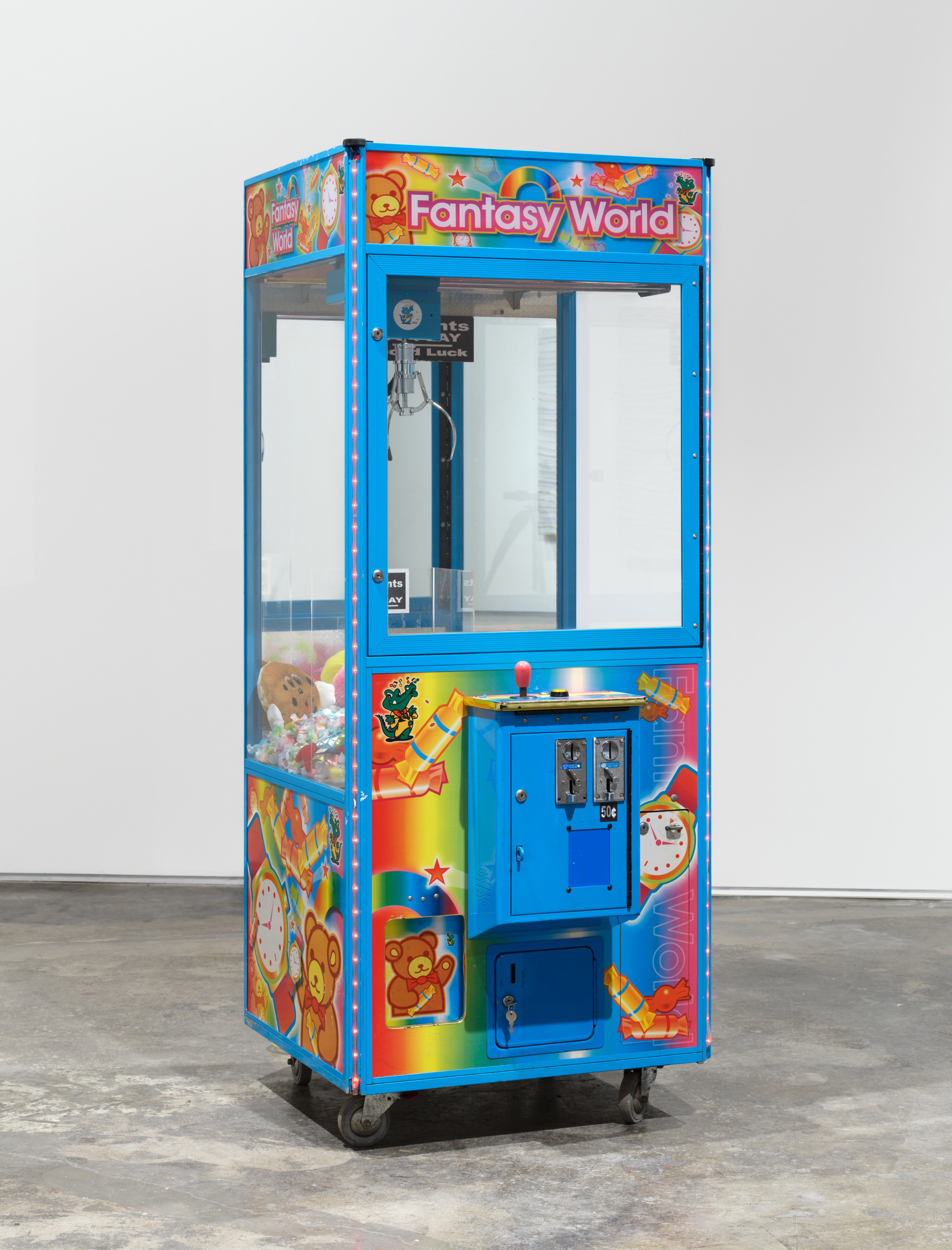 Anthony Olubunmi Akinbola artwork titled "Fantasy World". Blue carnival machine filled with saltwater taffy, rolls of cash and other toys. 75 3/4 x 31 x 34 1/4 in (192.4 x 78.7 x 87 cm), claw machine, various toys, and candy, 2023.